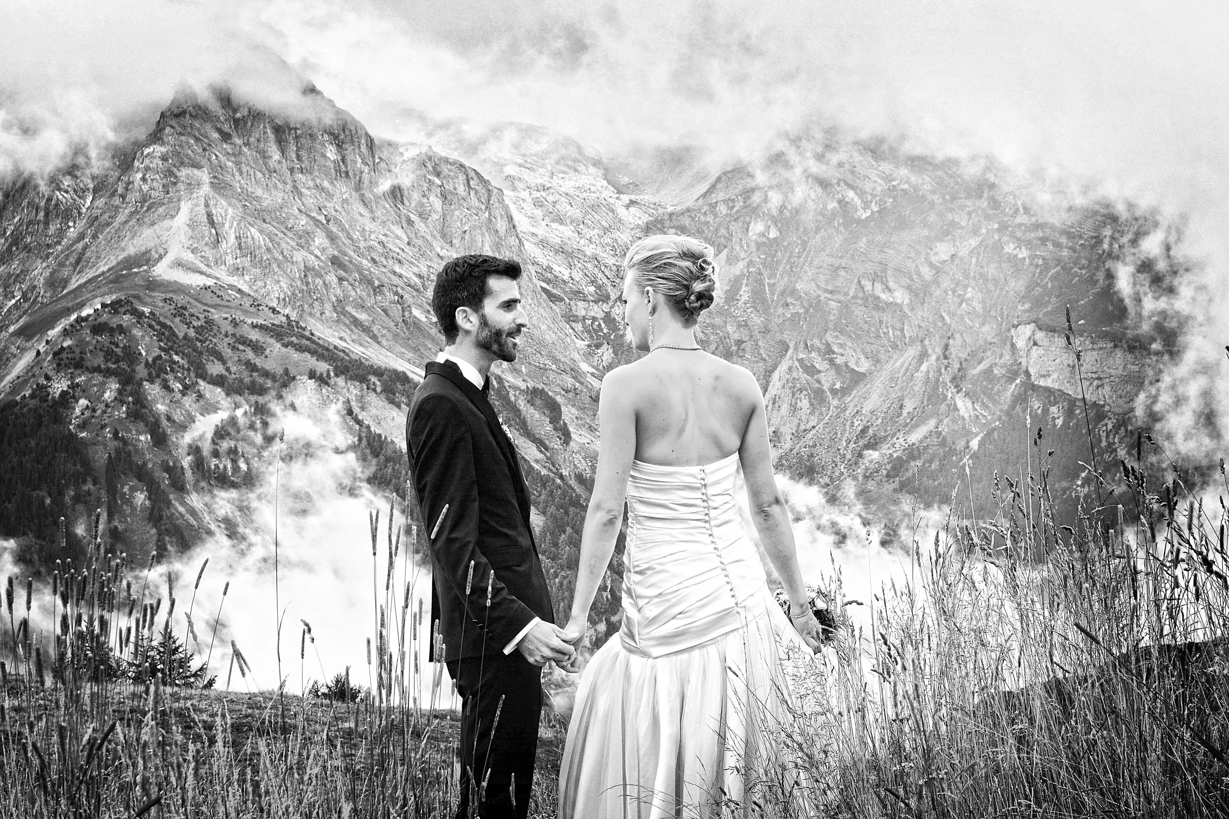 Un couple de mariés fait un vœu de mariage dans un paysage montagneux brumeux, l'homme porte un costume noir et la femme une robe de mariée blanche.