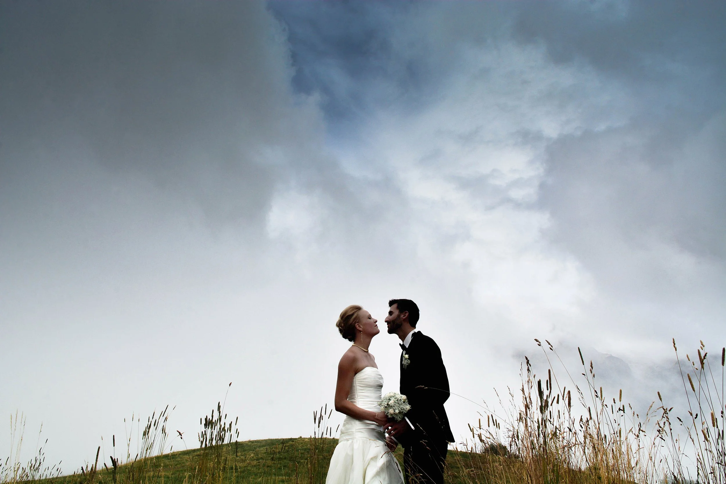 Un couple de mariés, une femme en robe blanche et un homme en costume noir, se tient sur une colline avec de l'herbe, sous un ciel orageux, partageant un moment intime avec des nuages sombres en arrière-plan.