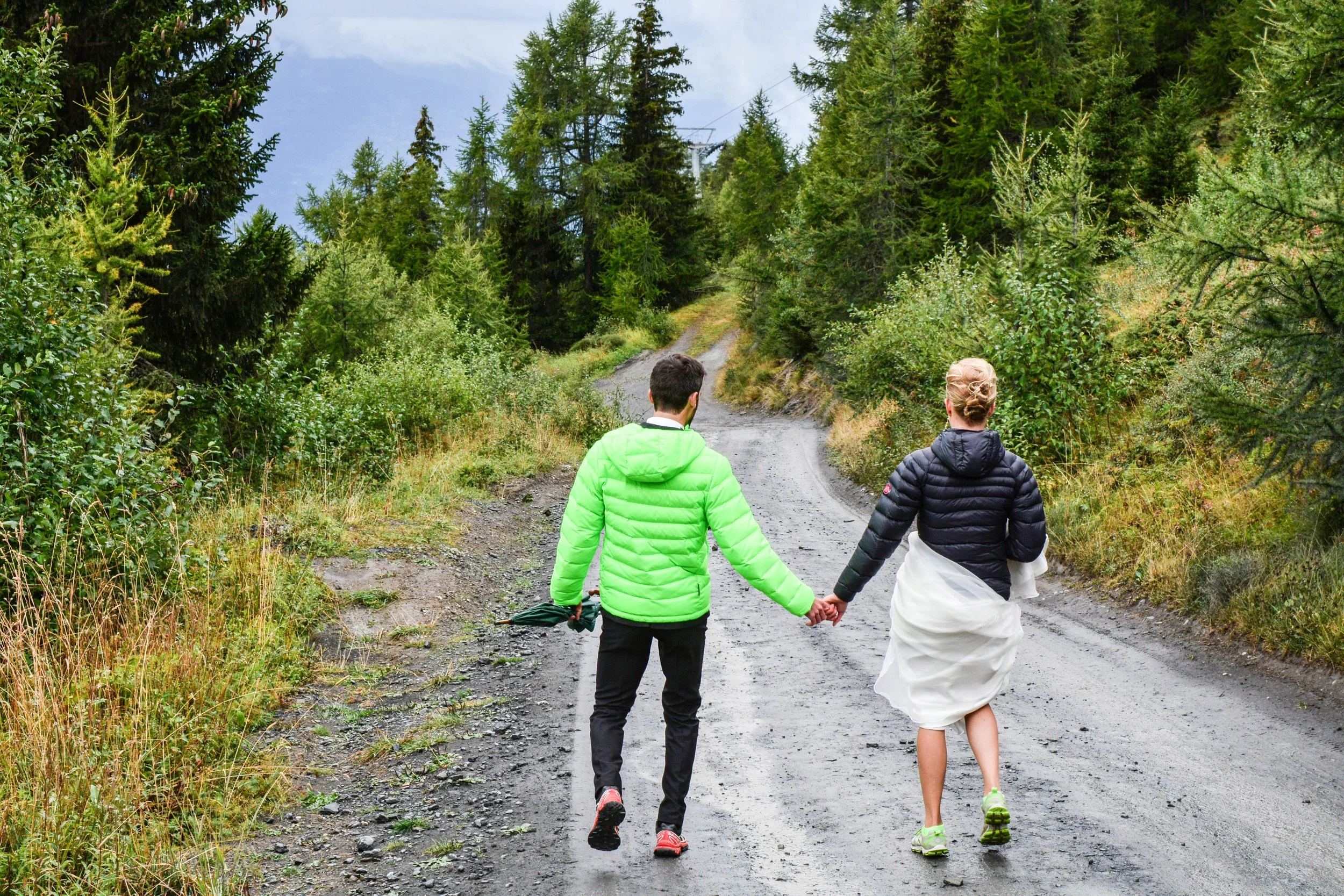 Deux personnes, un homme et une femme, marchent main dans la main sur un chemin de forêt, vêtus de vestes rembourrées, avec des arbres verts et un ciel nuageux en arrière-plan.