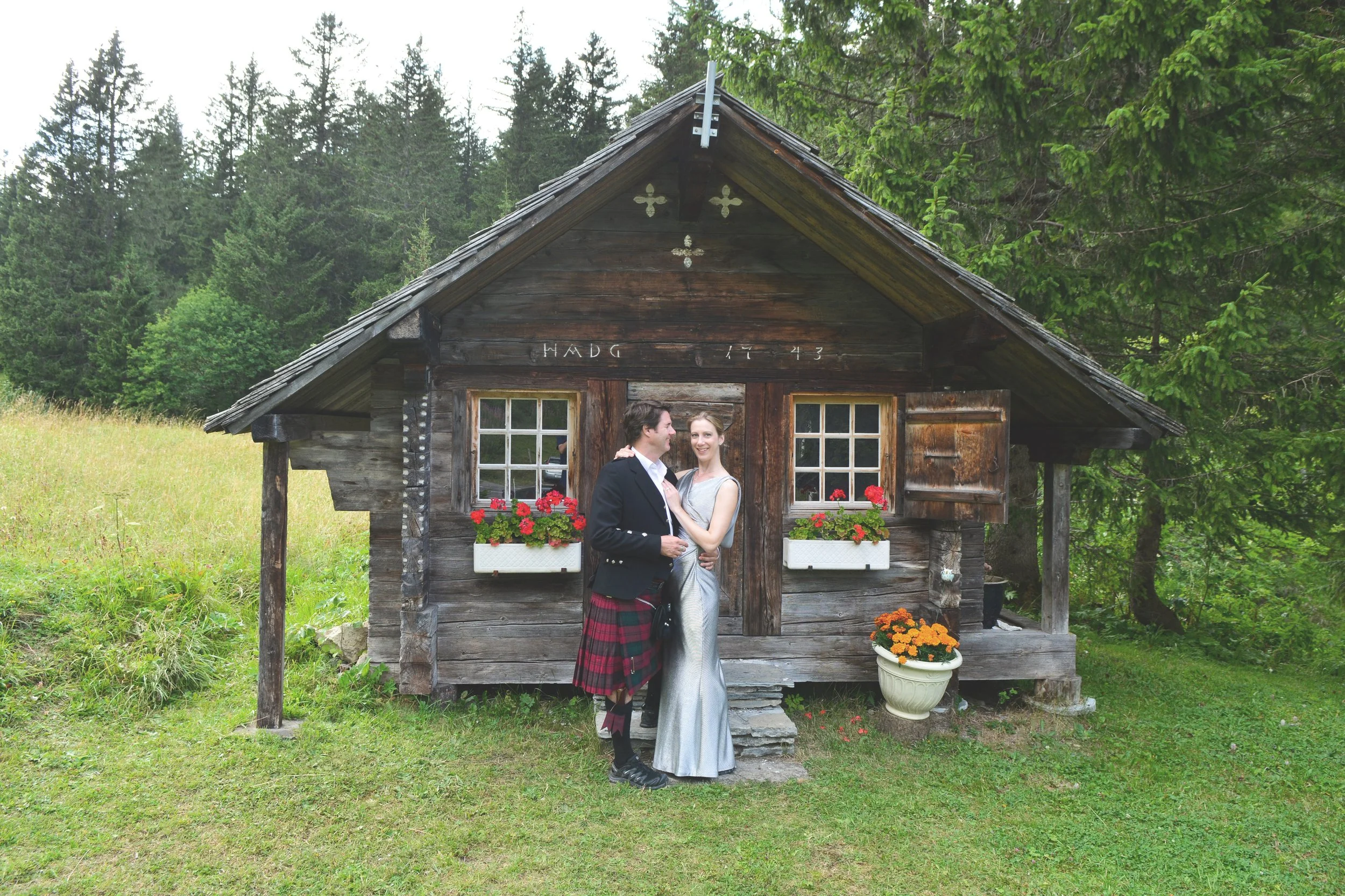 Un couple habillé élégamment devant une petite cabane en bois avec des fenêtres ornées de fleurs rouges, entourée de nature verdoyante.