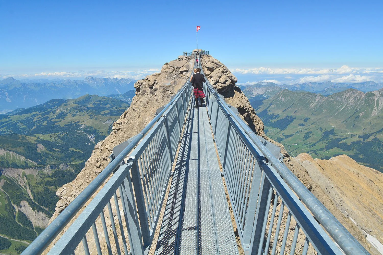 Une personne marche sur un pont en métal suspendu au sommet d'une montagne, avec un paysage de montagnes vertes et un ciel bleu en arrière-plan.