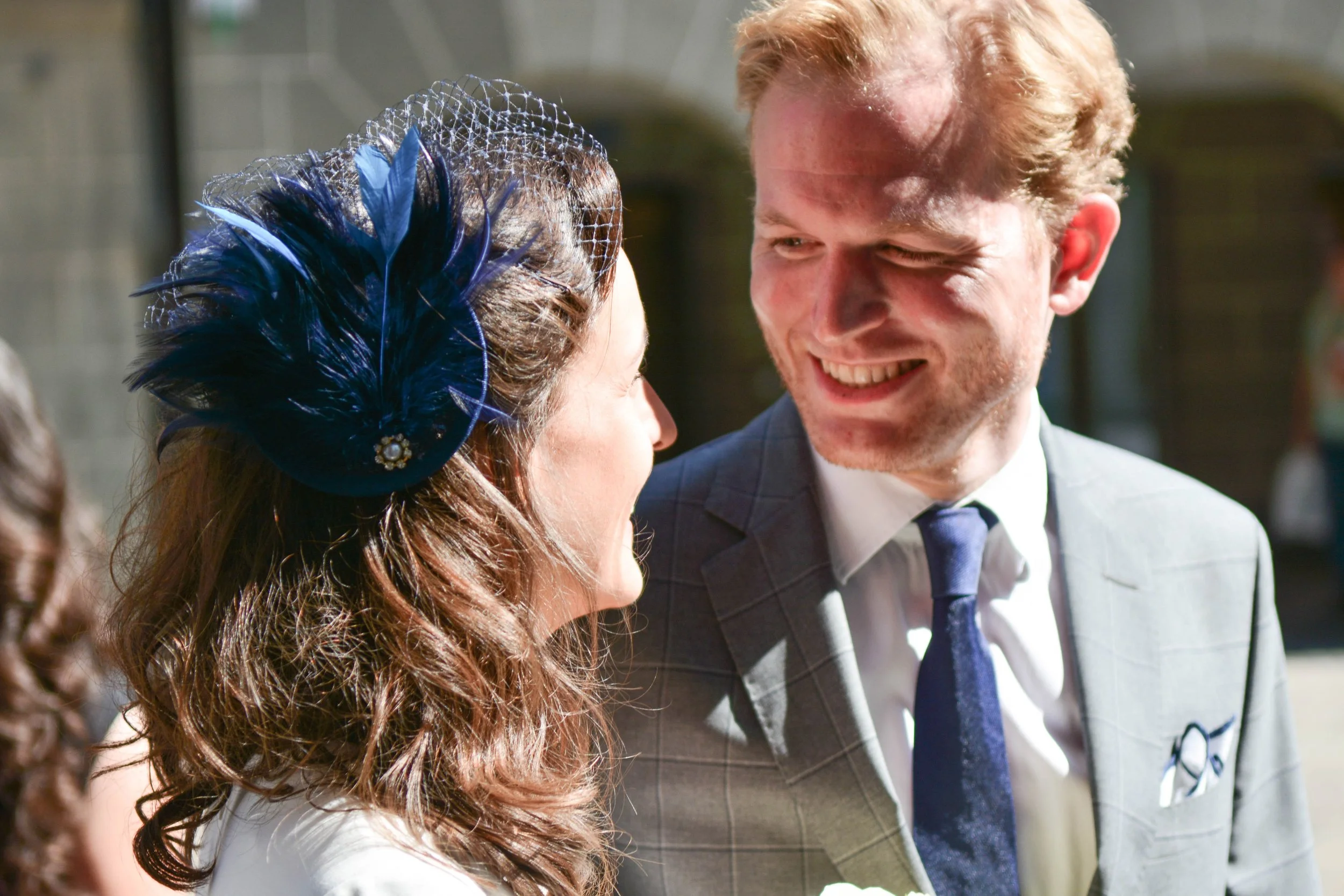 Un homme et une femme souriants bavardant lors d'un événement en plein air, la femme porte un accessoire en feathers bleu foncé avec voilette