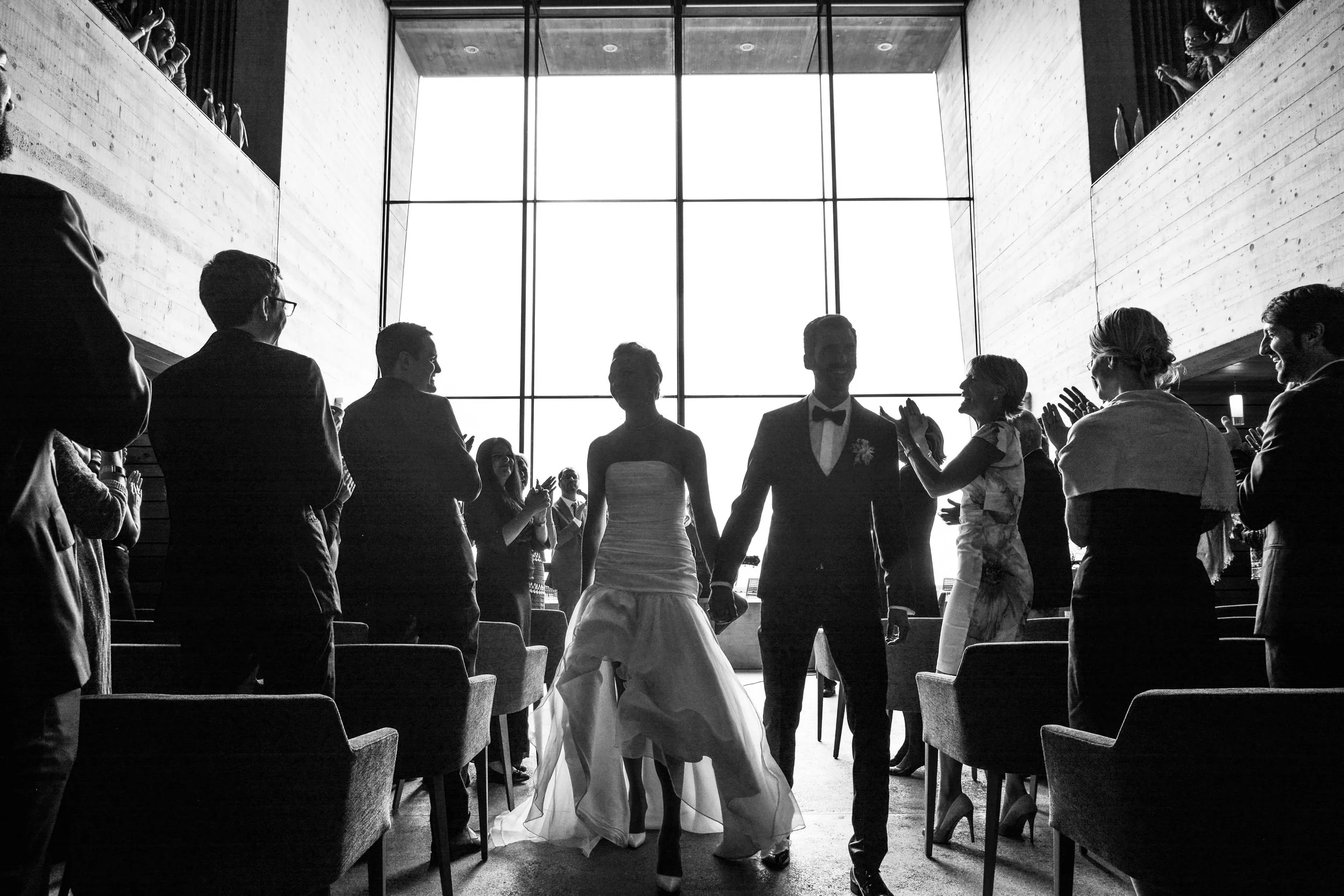 Un couple de mariés marche main dans la main dans une salle de mariage, entouré de leurs invités qui les applaudissent.