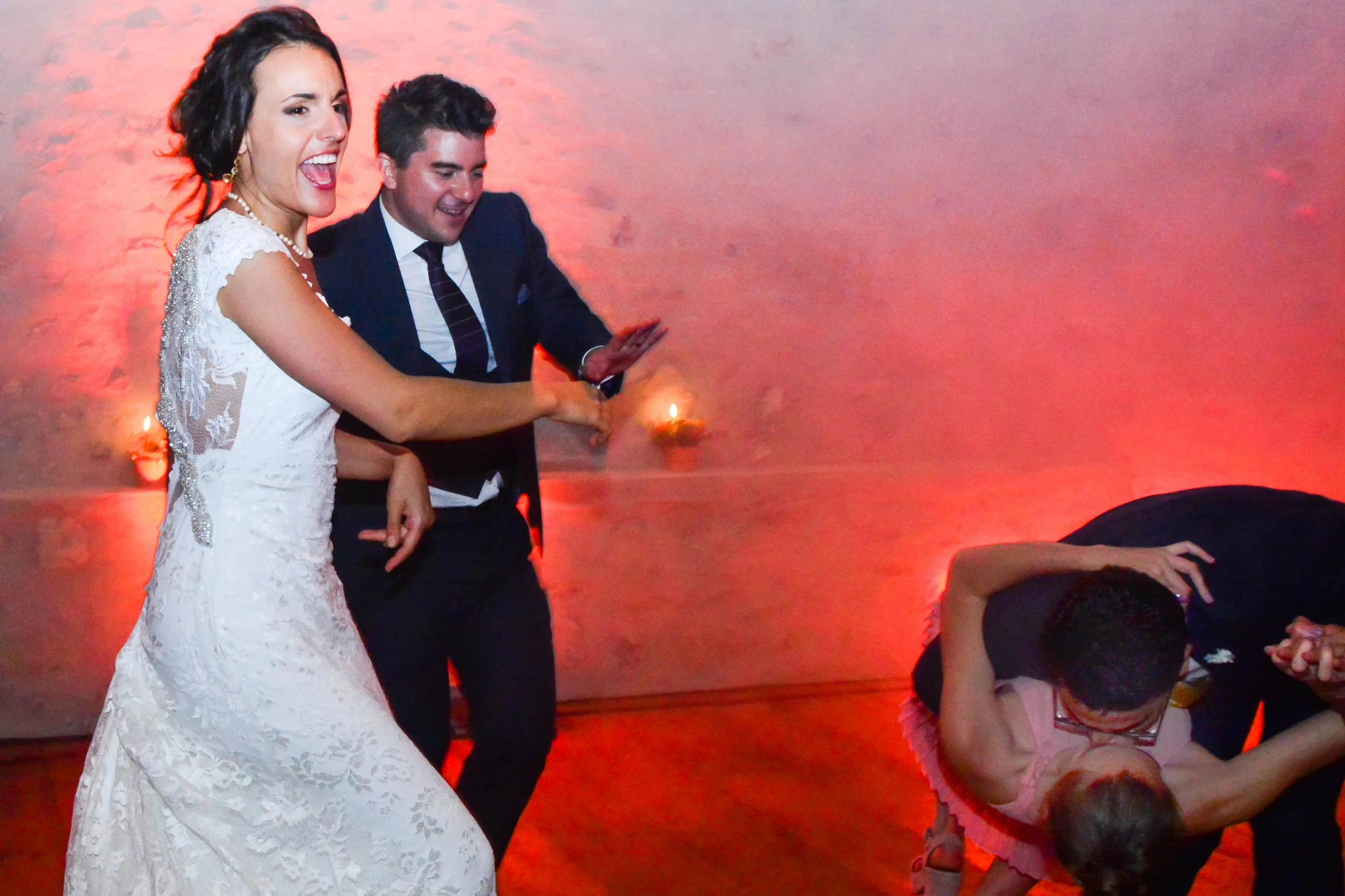 Un couple dansent lors d'un mariage, une femme en robe blanche et un homme en costume, avec un autre couple en arrière-plan, dans une ambiance chaleureuse avec un éclairage rouge.