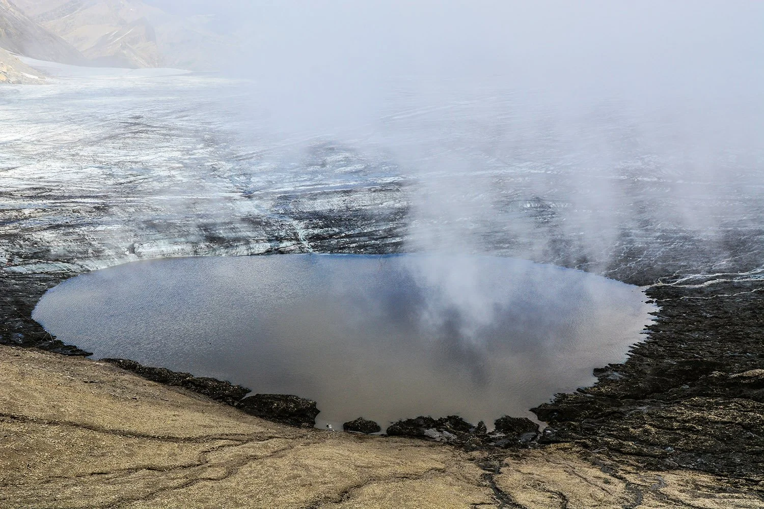 Un cratère volcanique rempli d'eau avec de la vapeur s'échappant, entouré de roches volcaniques.