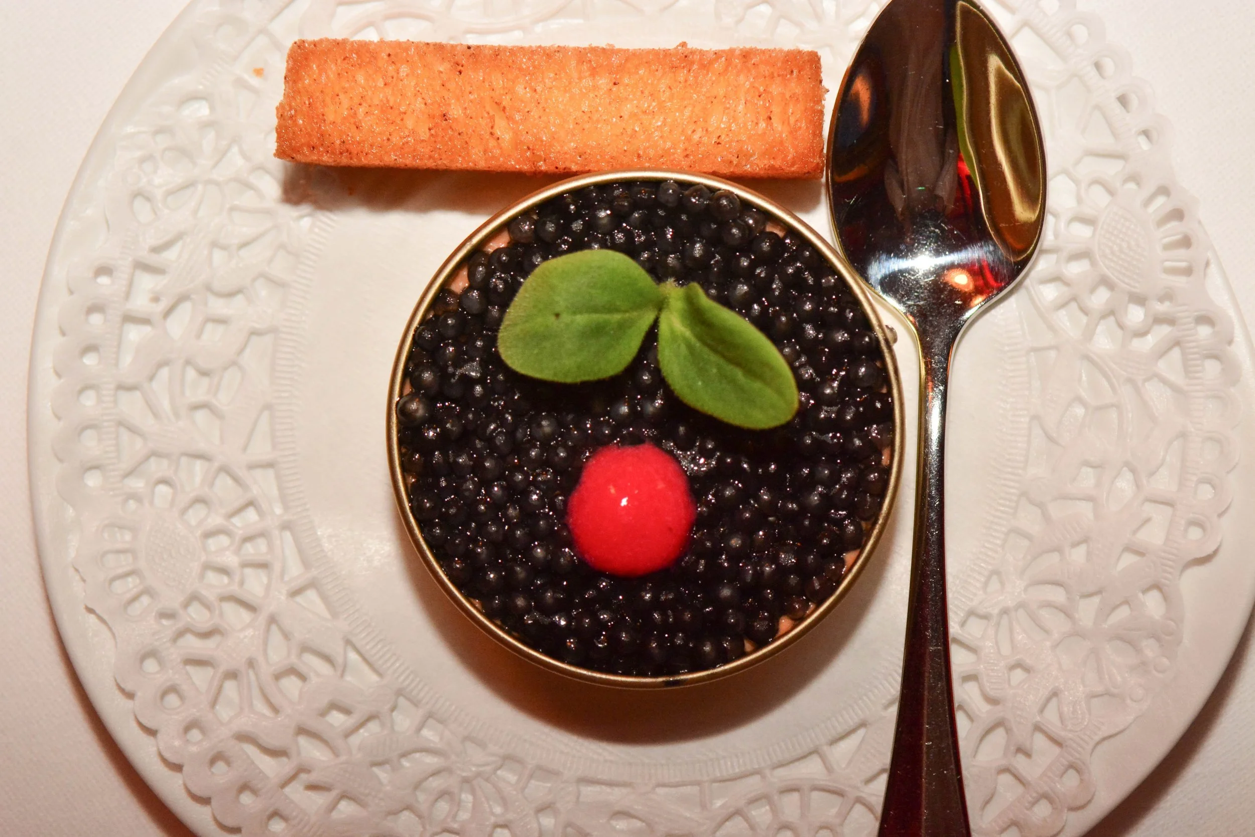 Dessert avec des petites baies noires, une feuille verte, une cerise rouge, une cuillère en argent, un biscuit rectangulaire croustillant, présenté sur une assiette blanche avec un motif décoratif