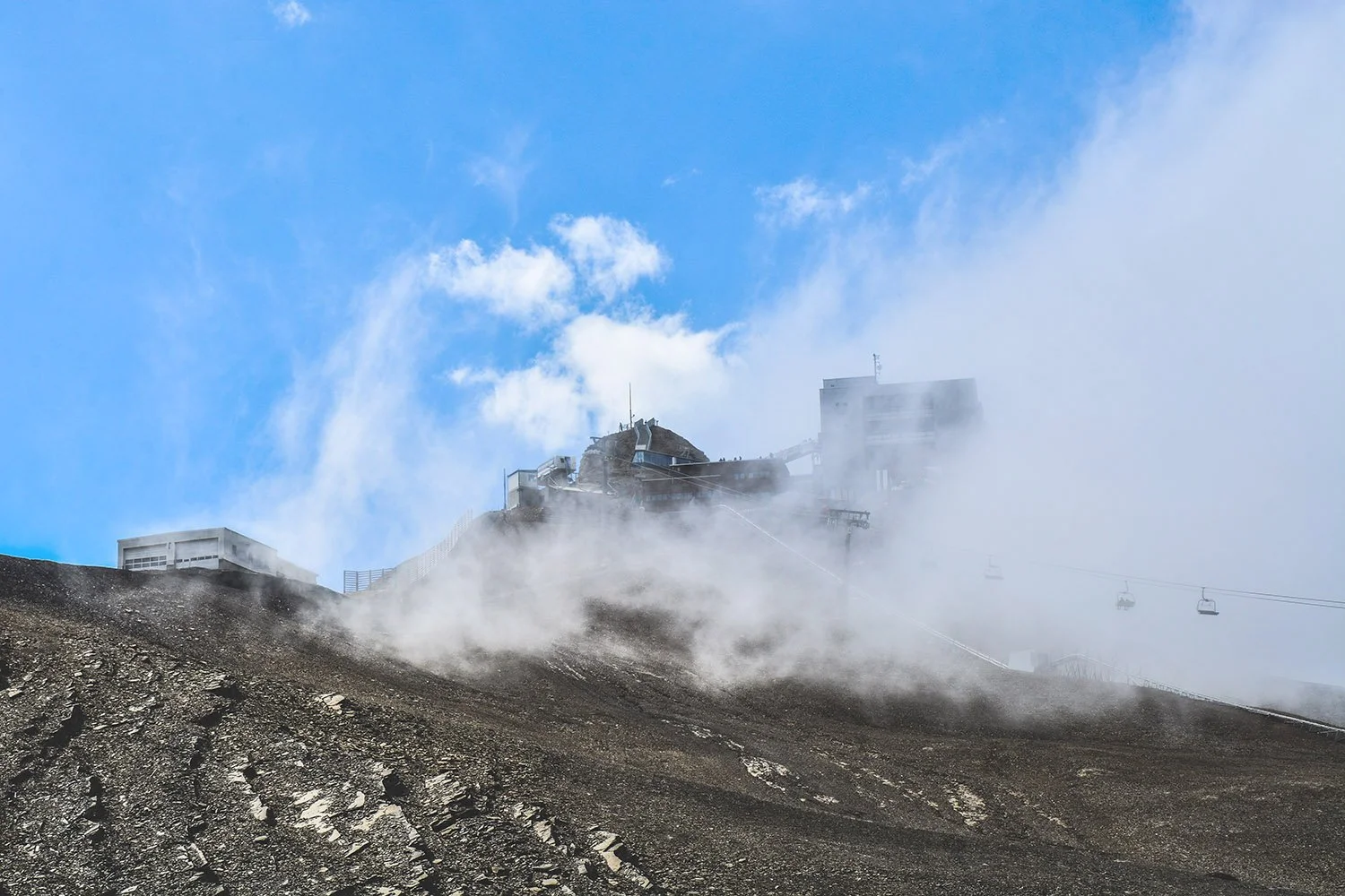 Vue d'une station de ski en haute montagne avec des bâtiments en hauteur, chaises de téléphérique, et de la vapeur ou de la fumée sur un terrain rocheux, avec un ciel bleu et quelques nuages.