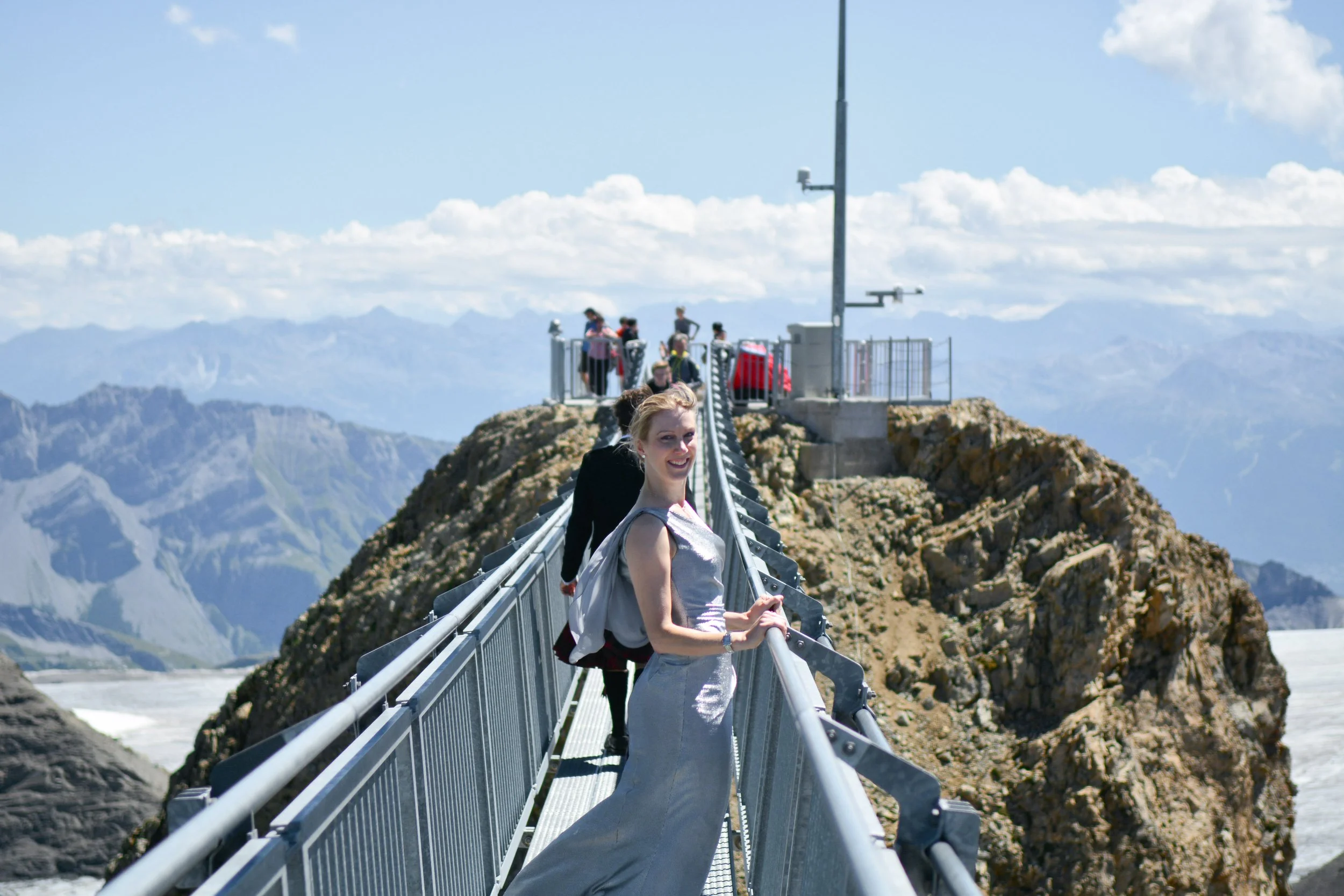 Une femme souriante portant une robe argentée et un sac à dos, debout sur un pont étroit suspendu en haut d'une montagne rocheuse, avec un groupe de touristes à l'arrière et un paysage de montagnes en arrière-plan.
