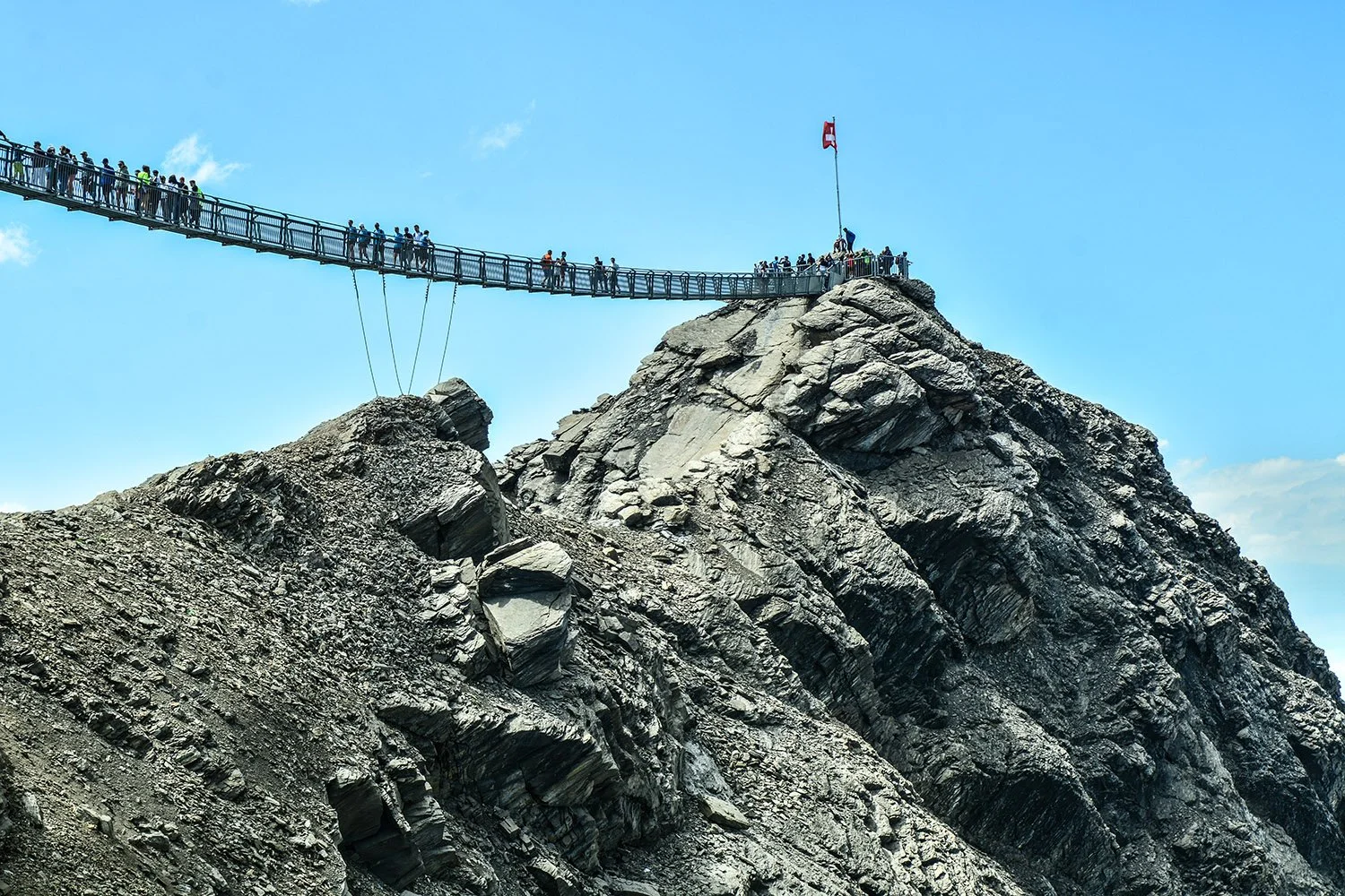 Un pont suspendu en acier traverse une montagne rocheuse, avec des groupes de personnes qui marchent dessus, sous un ciel bleu avec quelques nuages.