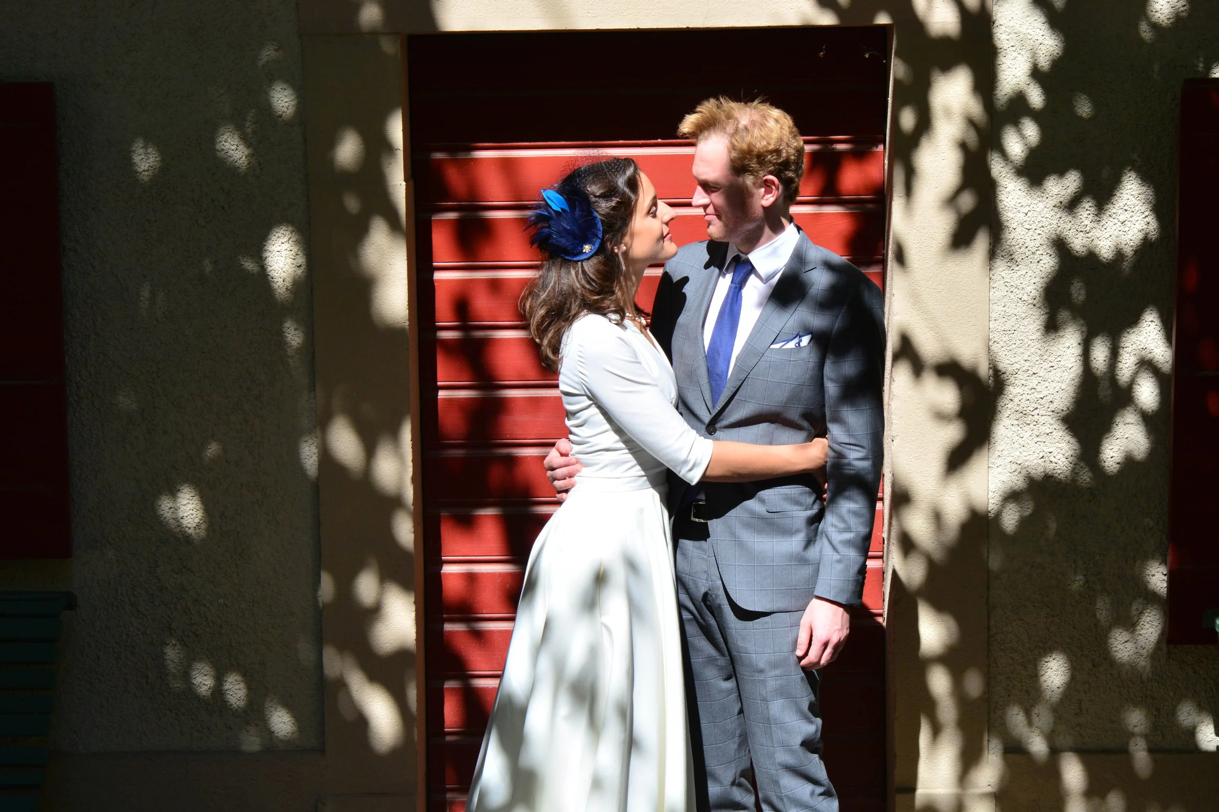 Un couple en habits élégants s'embrassant devant un mur avec des stores en bois rouges, entourés d'ombres de feuilles.
