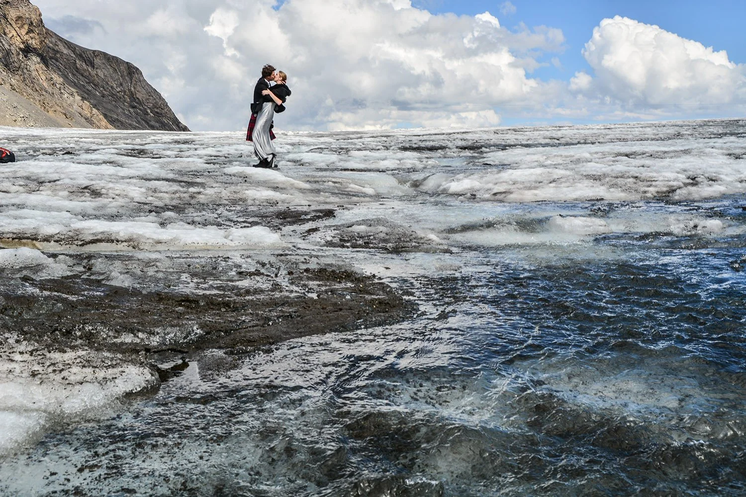 Deux personnes s'embrassent sur une surface de glace près de l'eau, avec un paysage de montagnes en arrière-plan et des nuages dans le ciel.
