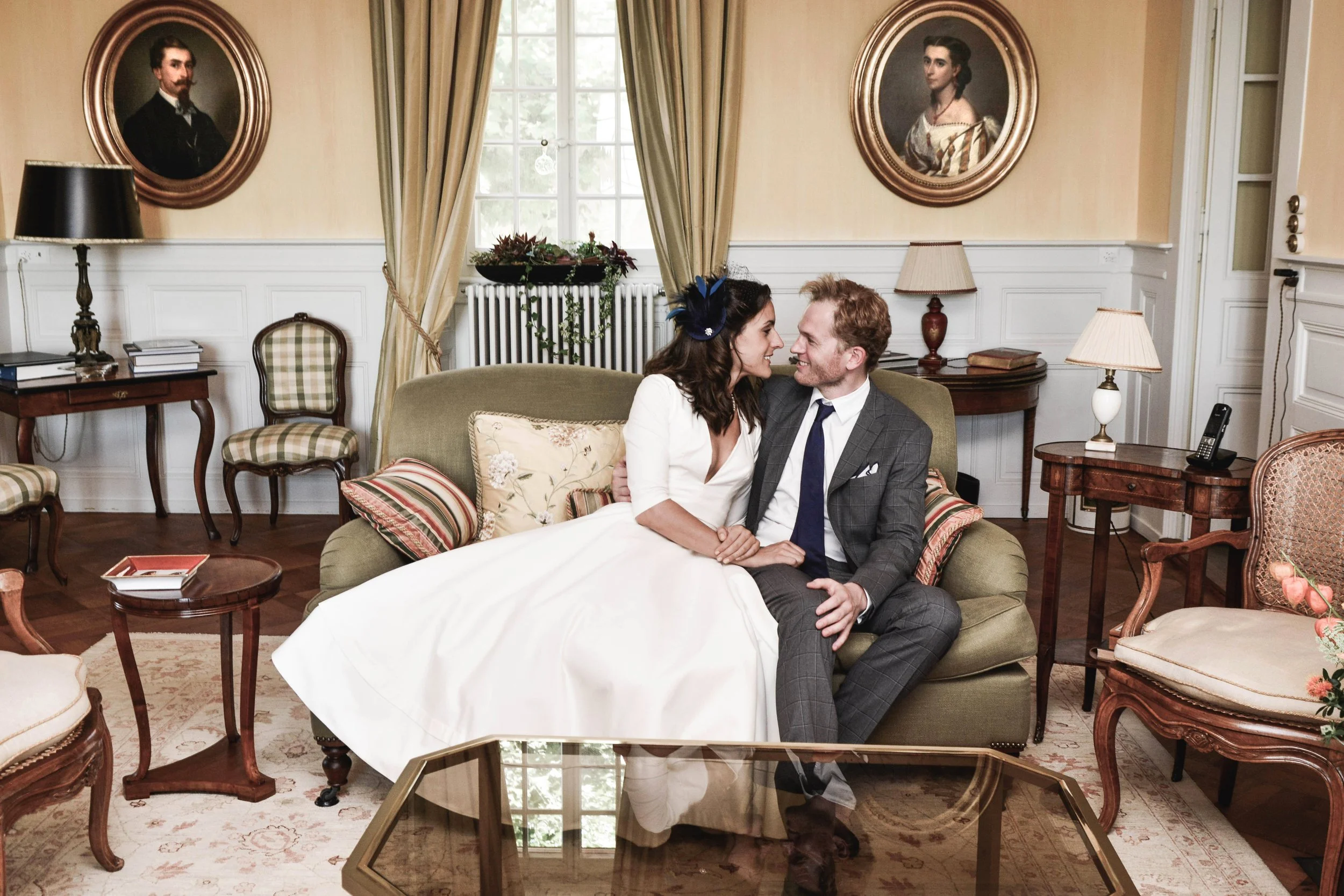 Un couple habillé formellement, une femme en robe blanche et un homme en costume gris, partage un moment intime sur un canapé dans un salon élégant avec des portraits à l'huile aux murs.