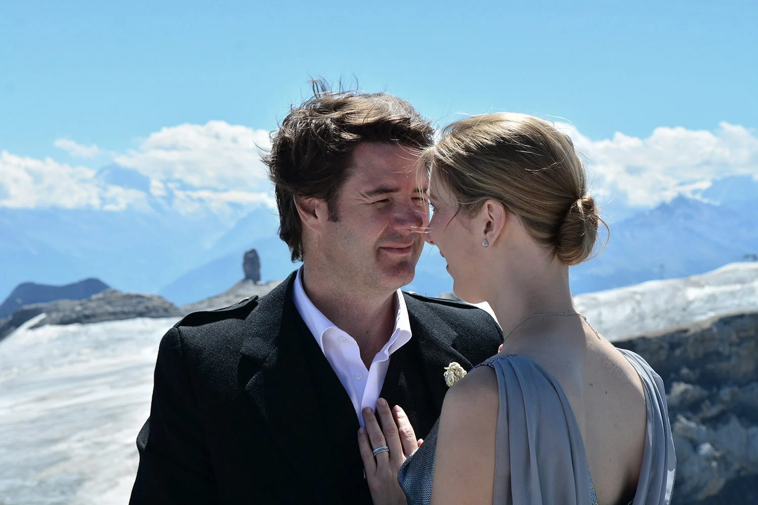 Un couple en vêtements de cérémonie, touchant leur front l'un contre l'autre, dans un décor de montagnes avec un glacier en arrière-plan, sous un ciel partiellement nuageux.