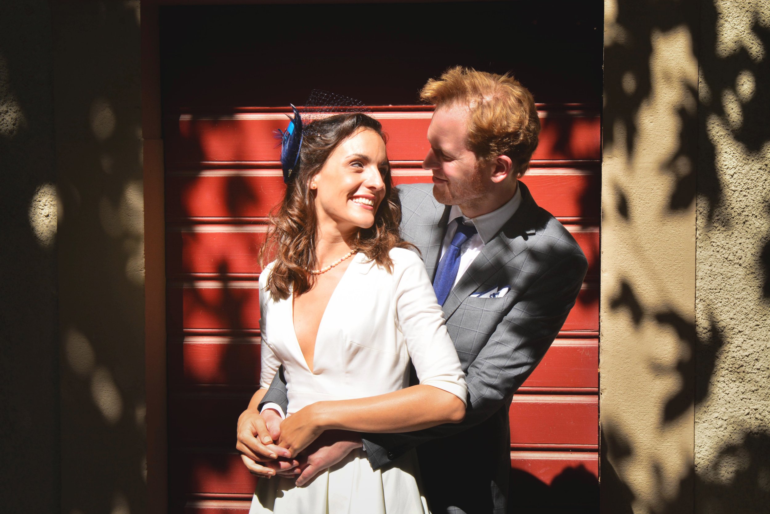 Un couple en costume souriant et se tenant dans les bras devant un mur en bois rouge, avec des ombres de branches d'arbre projetées sur le mur.