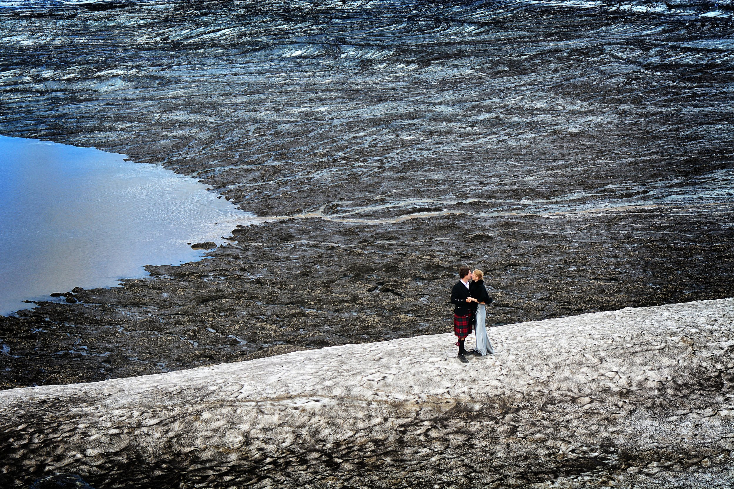 Deux personnes, un homme et une femme, se tiennent par la main sur un lit de neige au bord d'une étendue d'eau, dans un paysage naturel avec de la roche et de la glace.