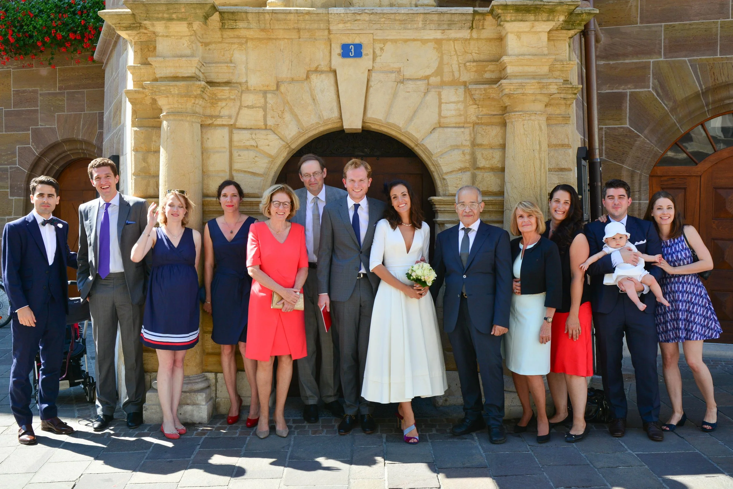 Groupe de personnes posant devant une entrée en pierre, probablement pour un mariage, avec des tenues formelles et une mariée tenant un bouquet de fleurs.