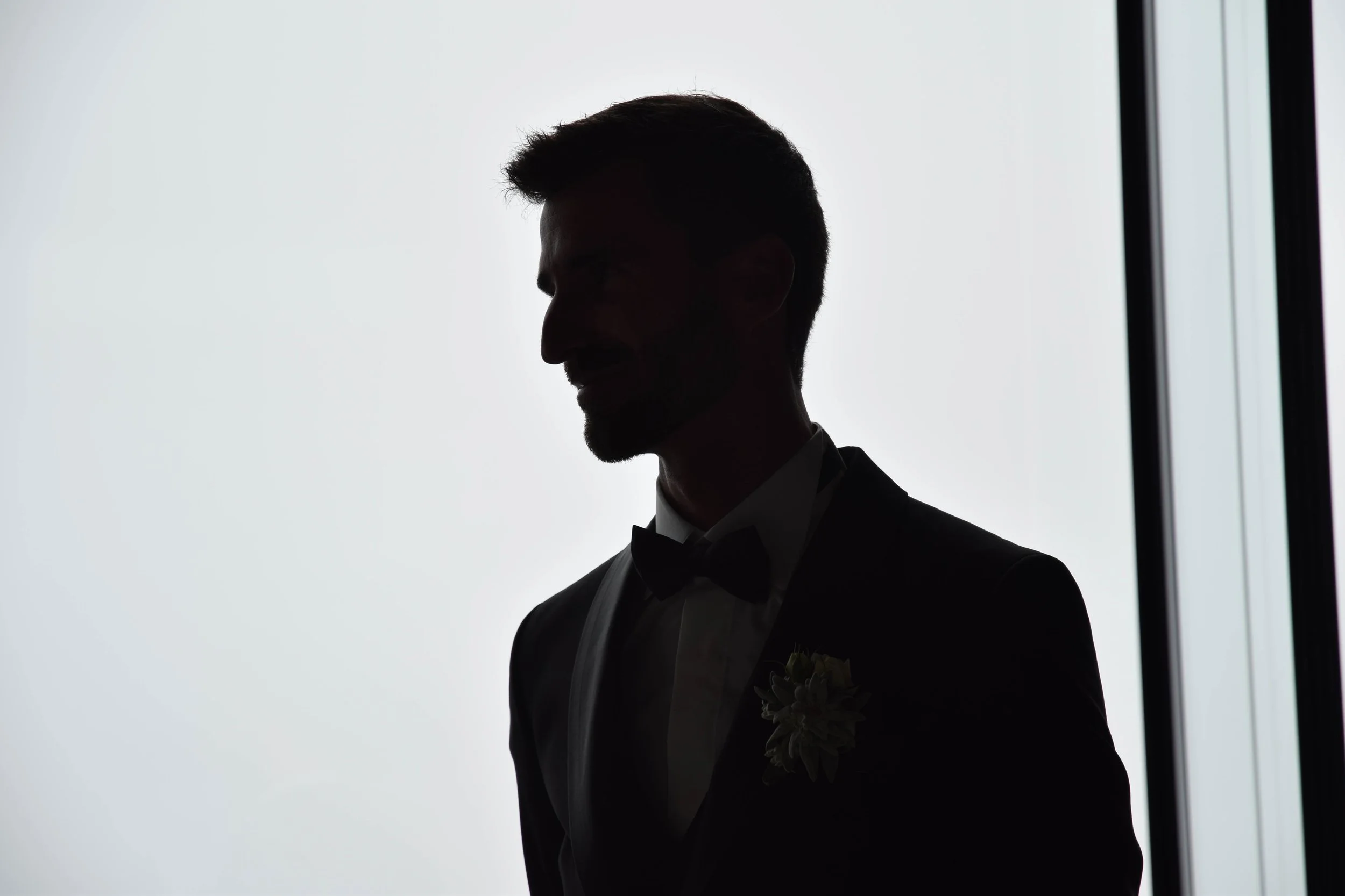 Silhouette d'un homme en costume de mariage avec un nœud papillon, portant une boutonnière, devant une lumière blanche, probablement à l'intérieur d'une pièce.