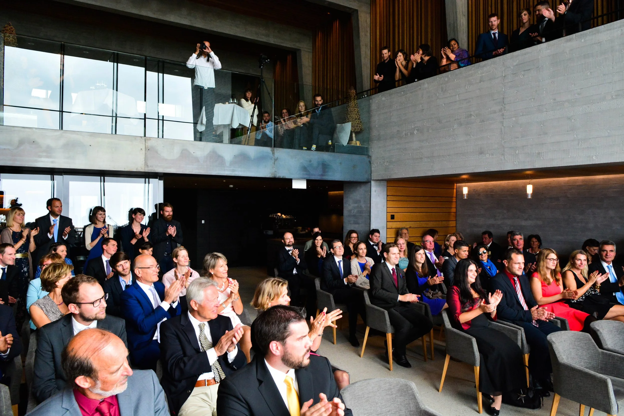 Une salle pleine de personnes assises et applaudissant lors d'un événement ou d'une cérémonie, avec un étage supérieur où d'autres personnes regardent et prennent des photos.