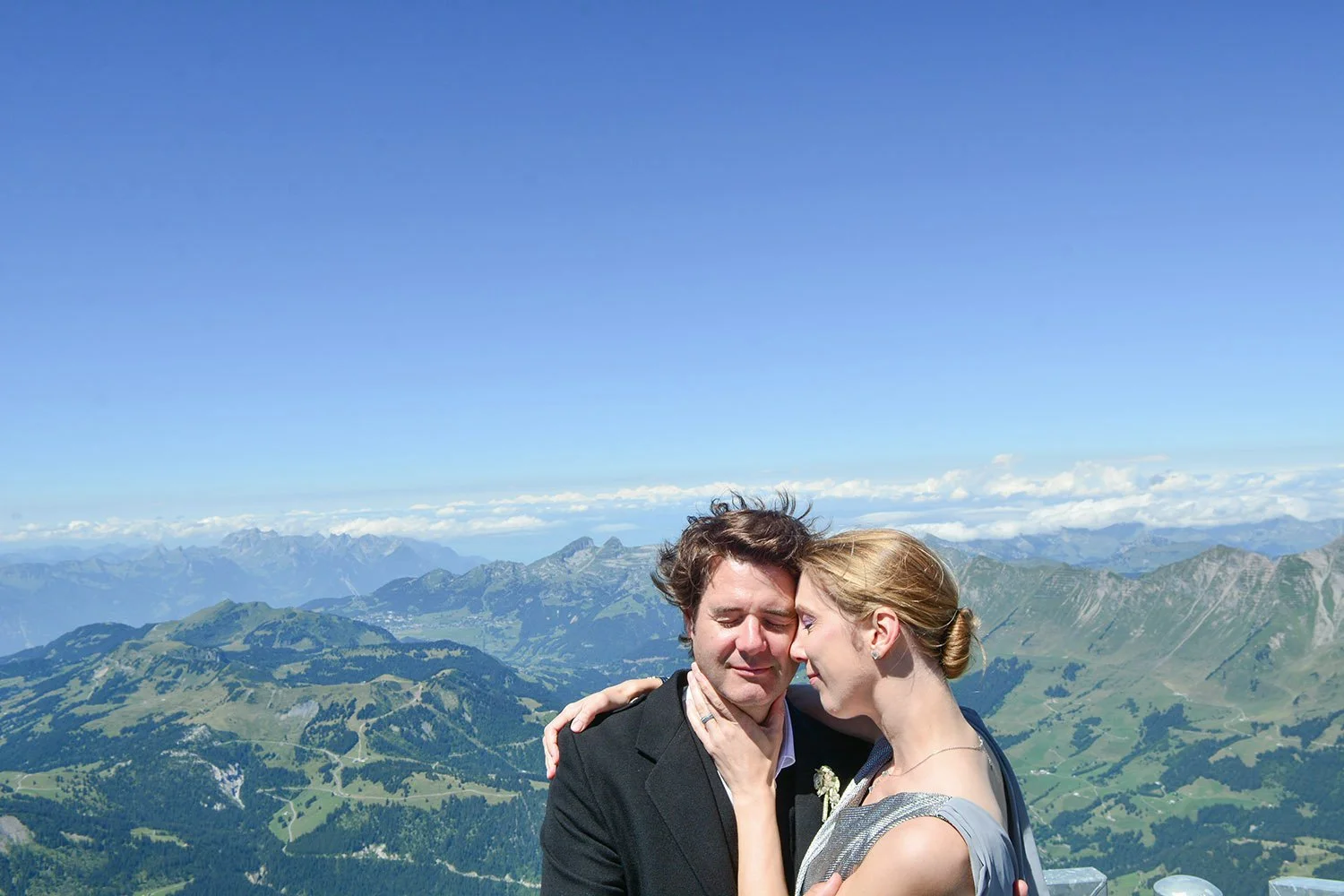 Un couple s'embrassant ou se tenant près du visage avec un paysage de montagnes en arrière-plan, ciel bleu clair.