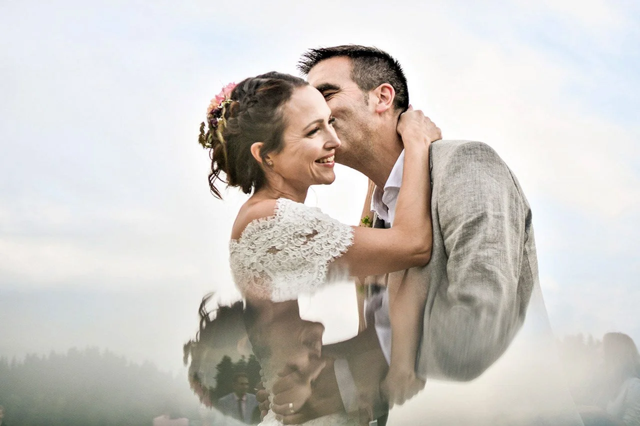 Un couple de mariés souriant, en plein baiser, lors de leur mariage en extérieur sous un ciel partiellement nuageux.
