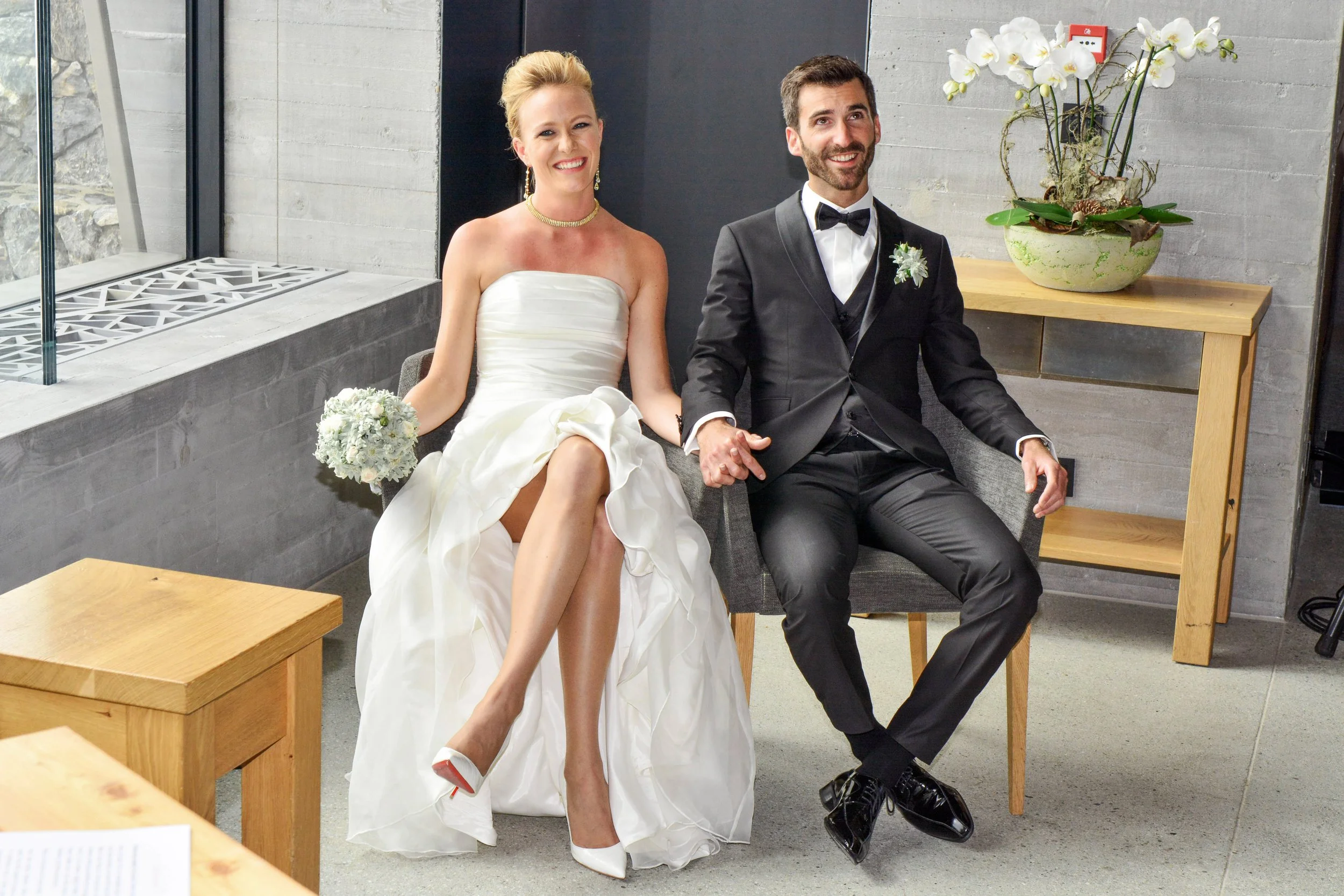 Un couple en costume de mariage assis dans une pièce moderne, la femme en robe de mariée blanche et le homme en costume noir avec un nœud papillon, se tenant la main et souriant.