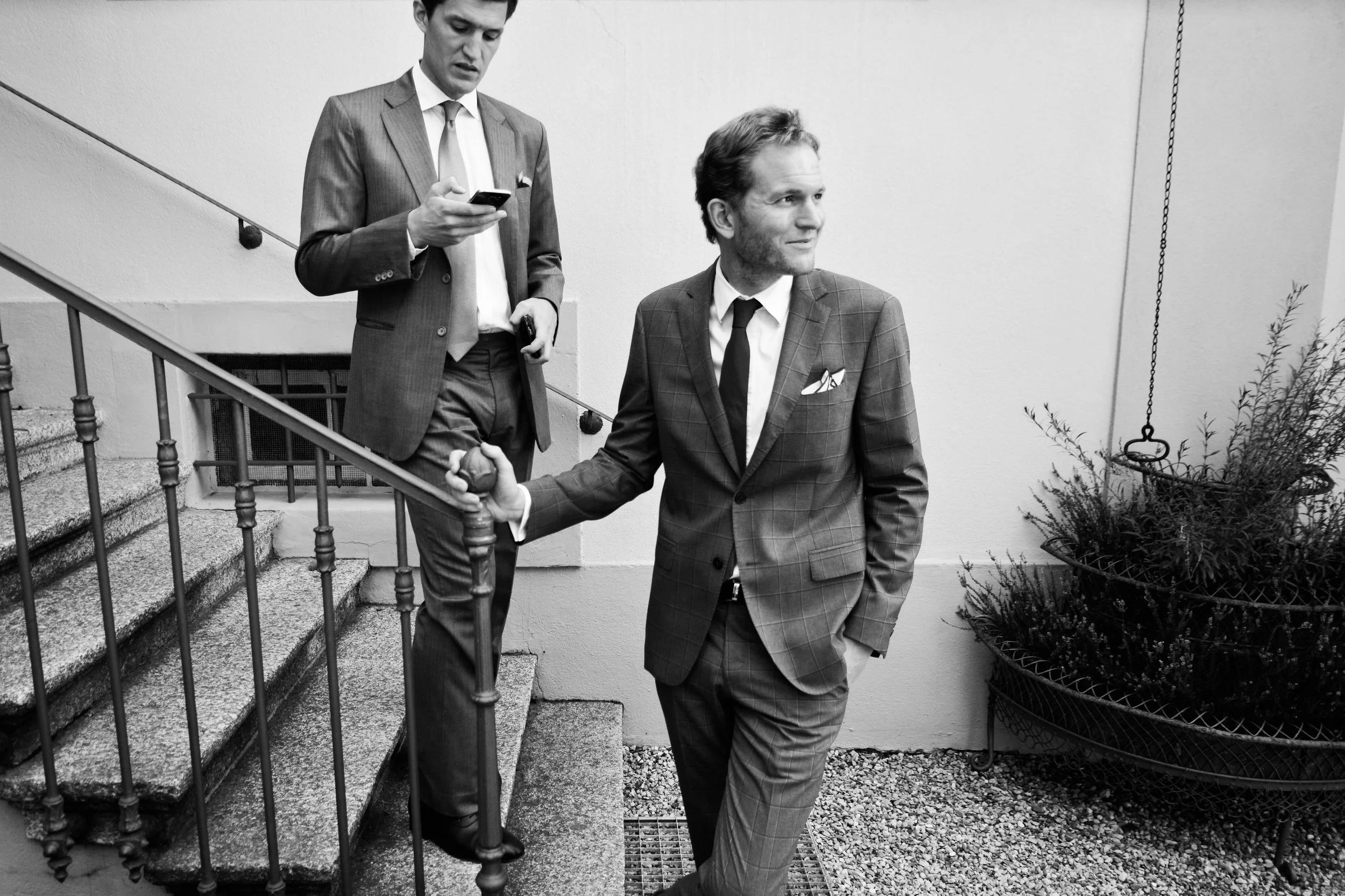 Deux hommes en costumes habillés élégamment, l'un descendant des escaliers en tenant une balle de tennis, l'autre debout à côté, regardant de côté, dans un environnement intérieur avec des plantes.