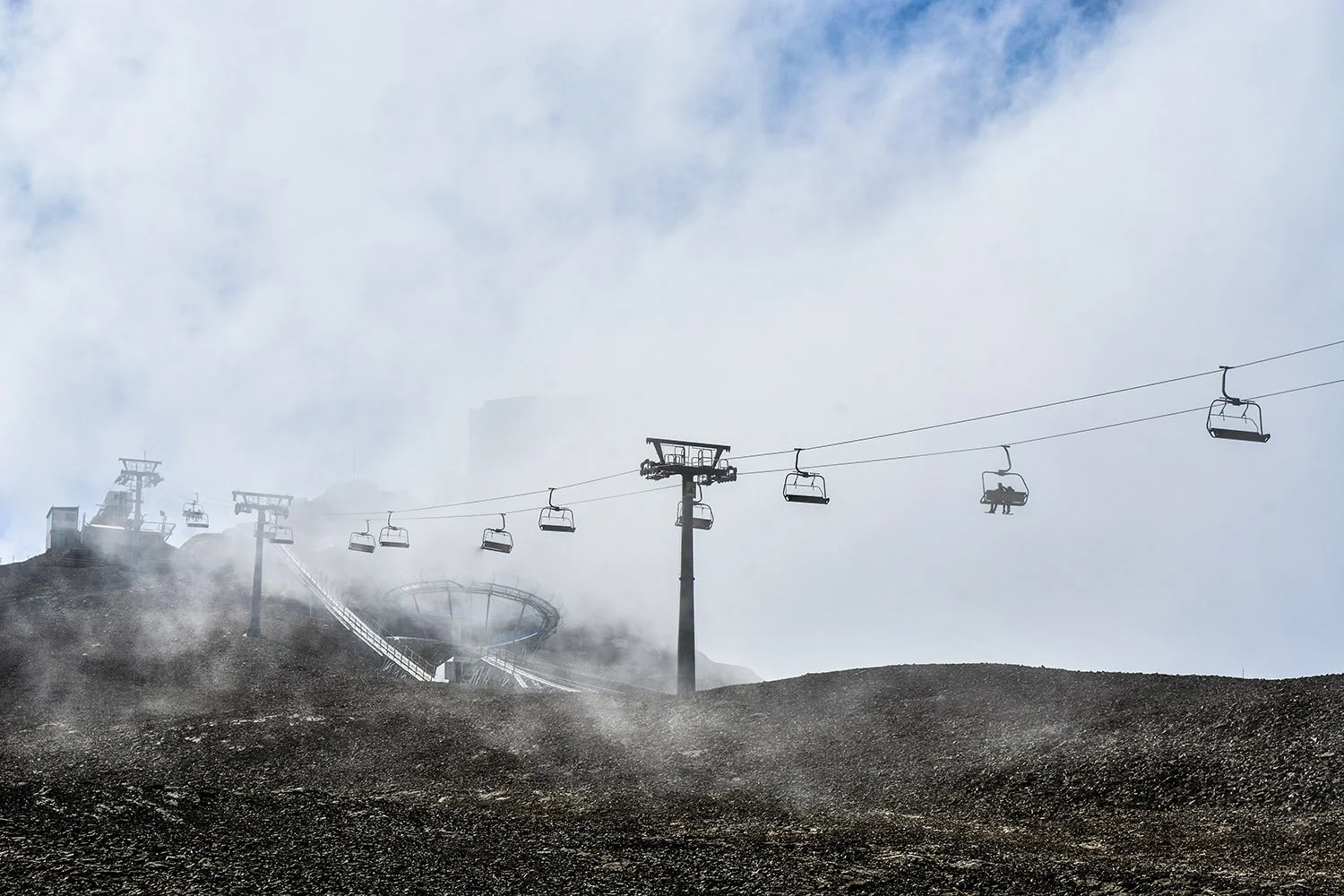 Chaises longues du téléphérique suspendues au-dessus d'une zone rocheuse dans la brume, avec un sommet de montagne derrière, sous un ciel nuageux.