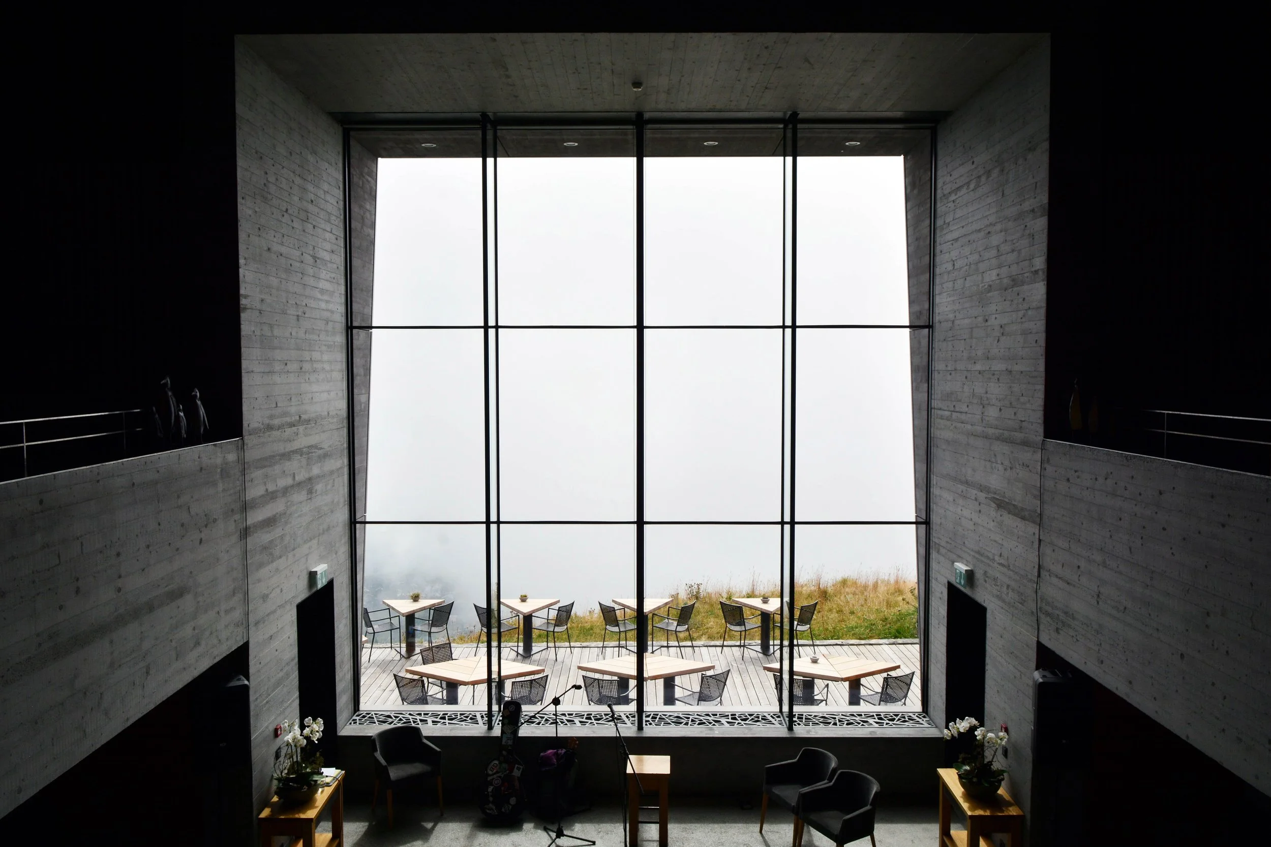 Vue intérieure d'un bâtiment moderne - Chetzeron - avec une grande fenêtre donnant sur une terrasse extérieure avec des tables et des chaises, murs en béton gris et décoration minimaliste.