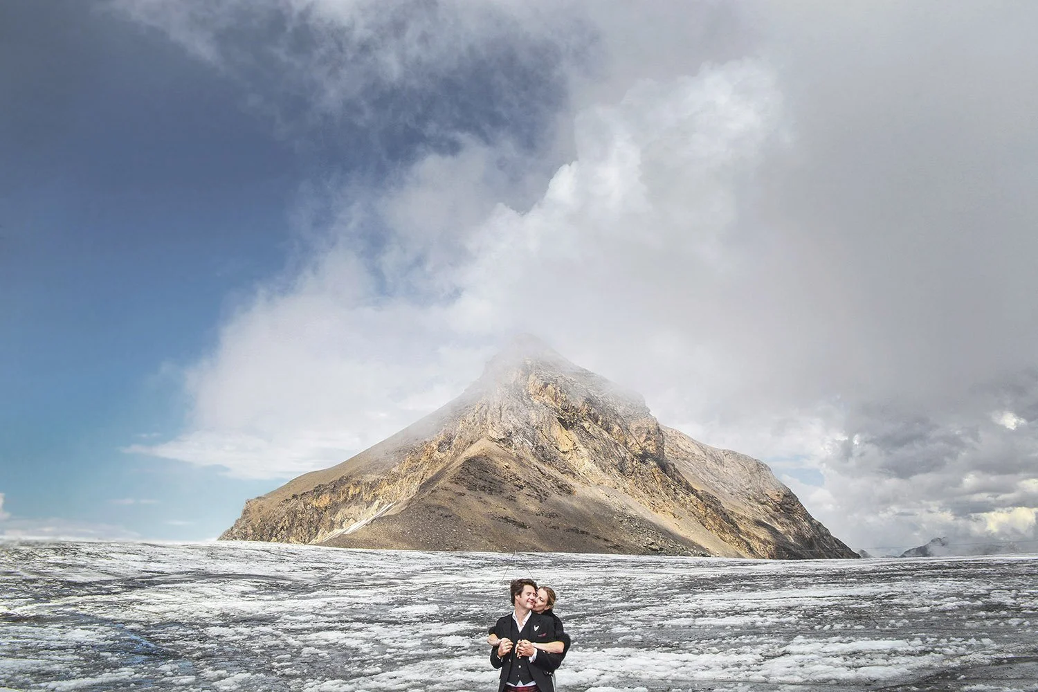 Deux personnes, un homme et une femme, se tiennent sur une surface glacée avec une montagne en arrière-plan, en partie couverte de nuages et de brouillard.