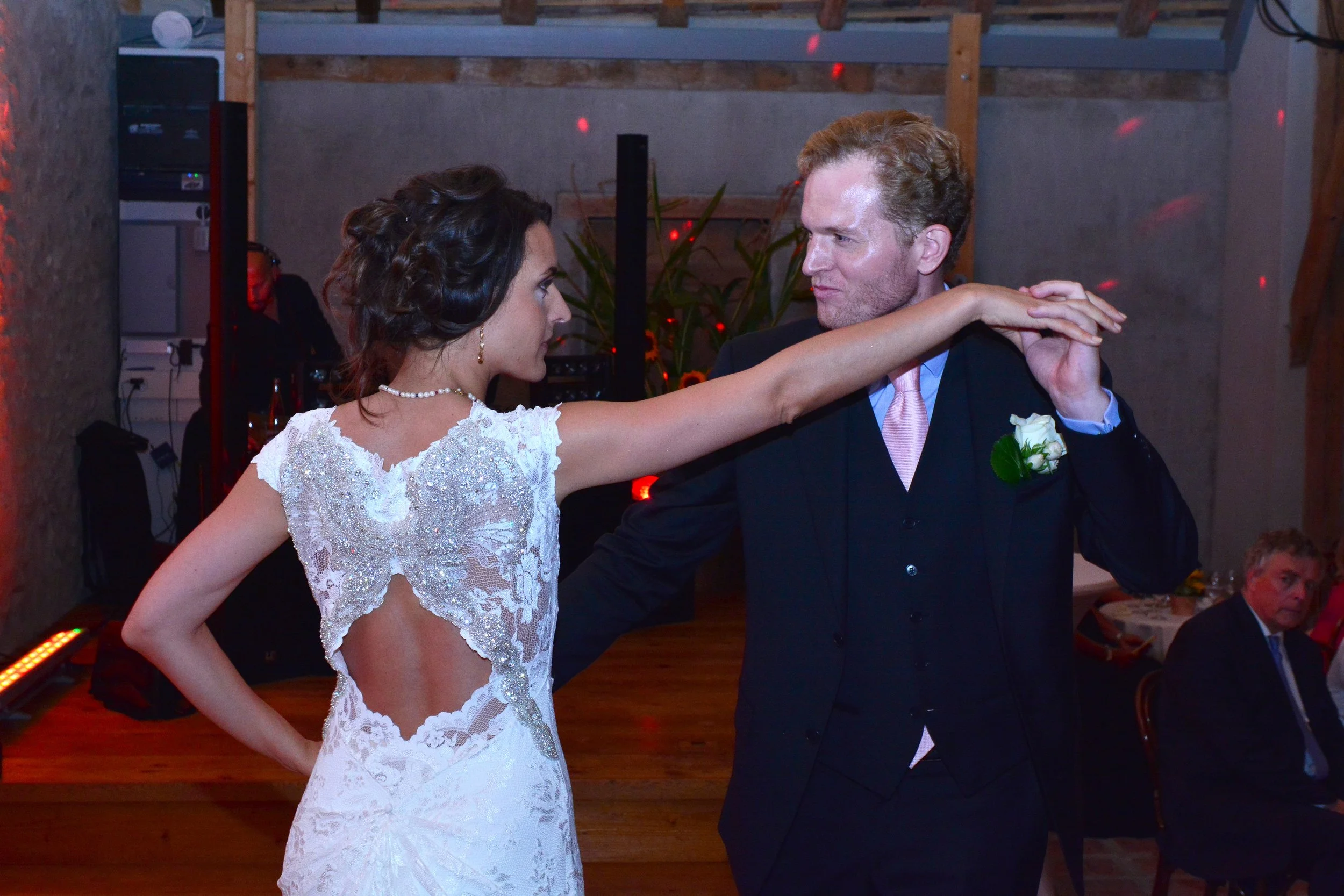 Un couple danse lors de leur mariage, la femme porte une robe blanche avec des détails en dentelle et un homme en costume noir avec une boutonnière à une fête.