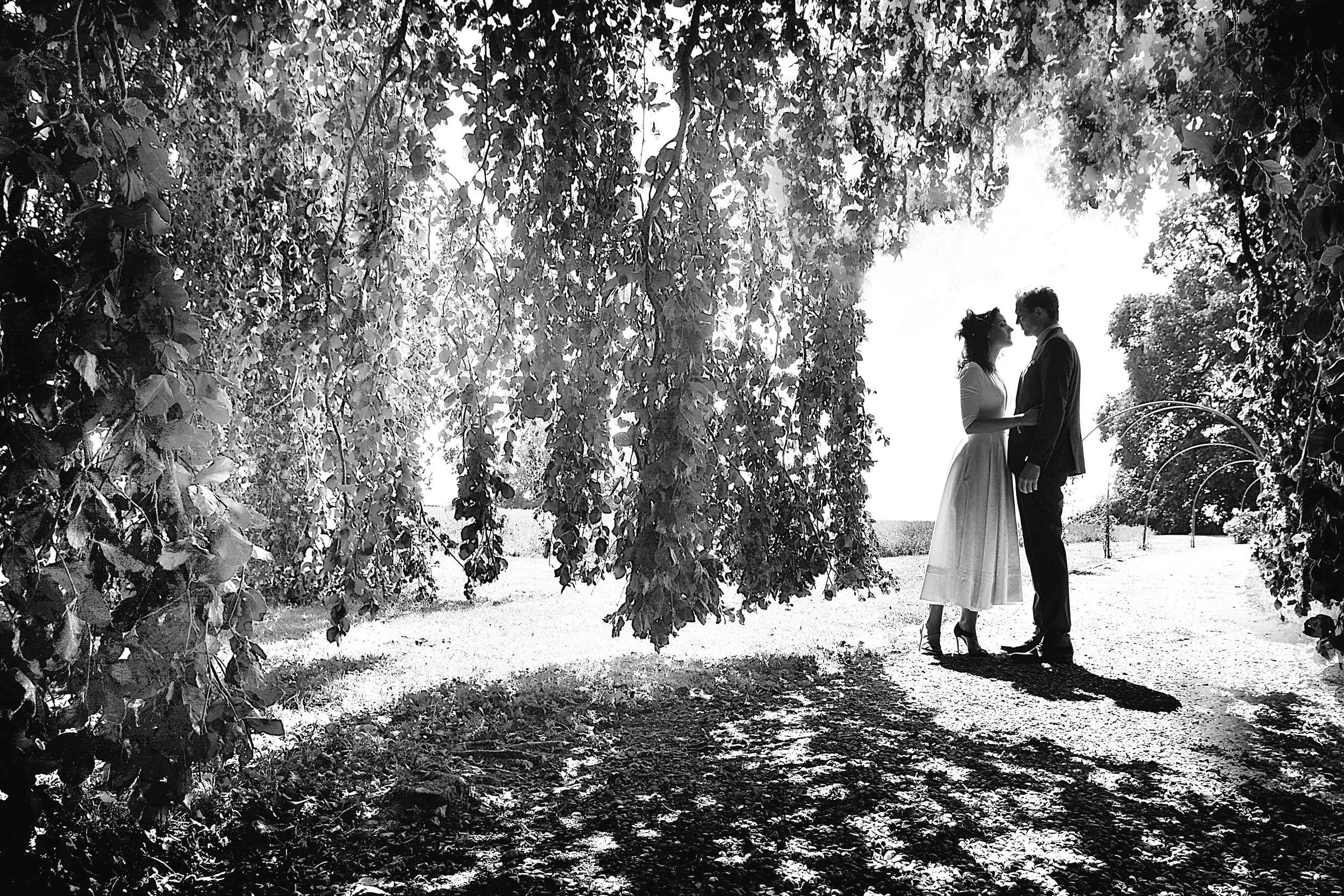 Deux personnes qui semblent être un couple se tiennent par la main sous une arche fleurie en plein air, en scène en noir et blanc.