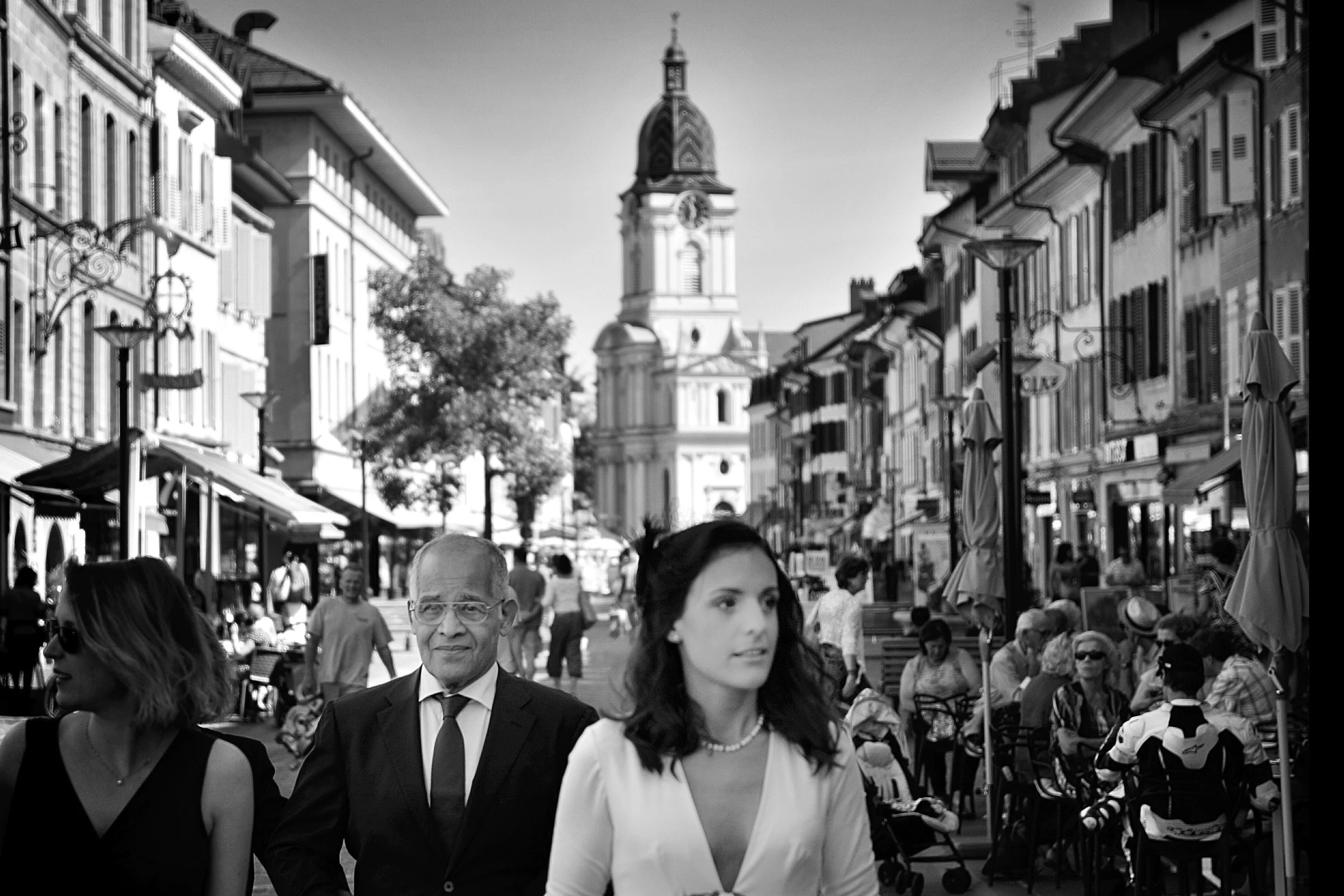 Une scène urbaine animée avec des personnes qui se promènent dans une rue piétonne, des cafés en plein air et une église avec un clocher en arrière-plan.