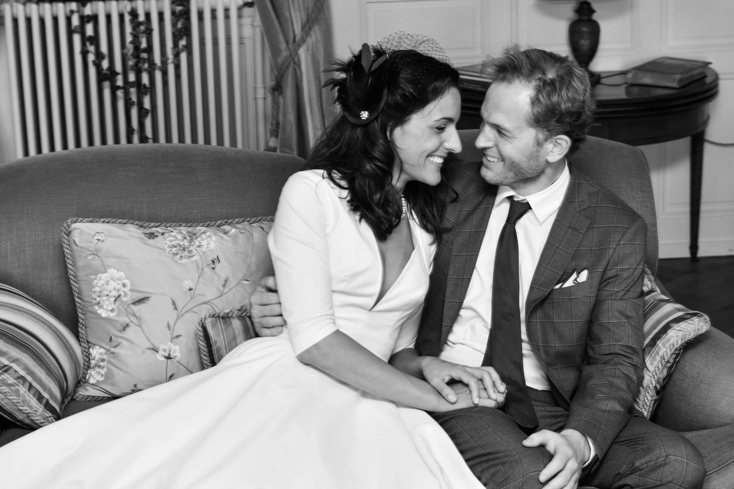 Un couple souriant, habillé élégamment, assis sur un canapé dans un intérieur chic, échangeant un regard affectueux.
