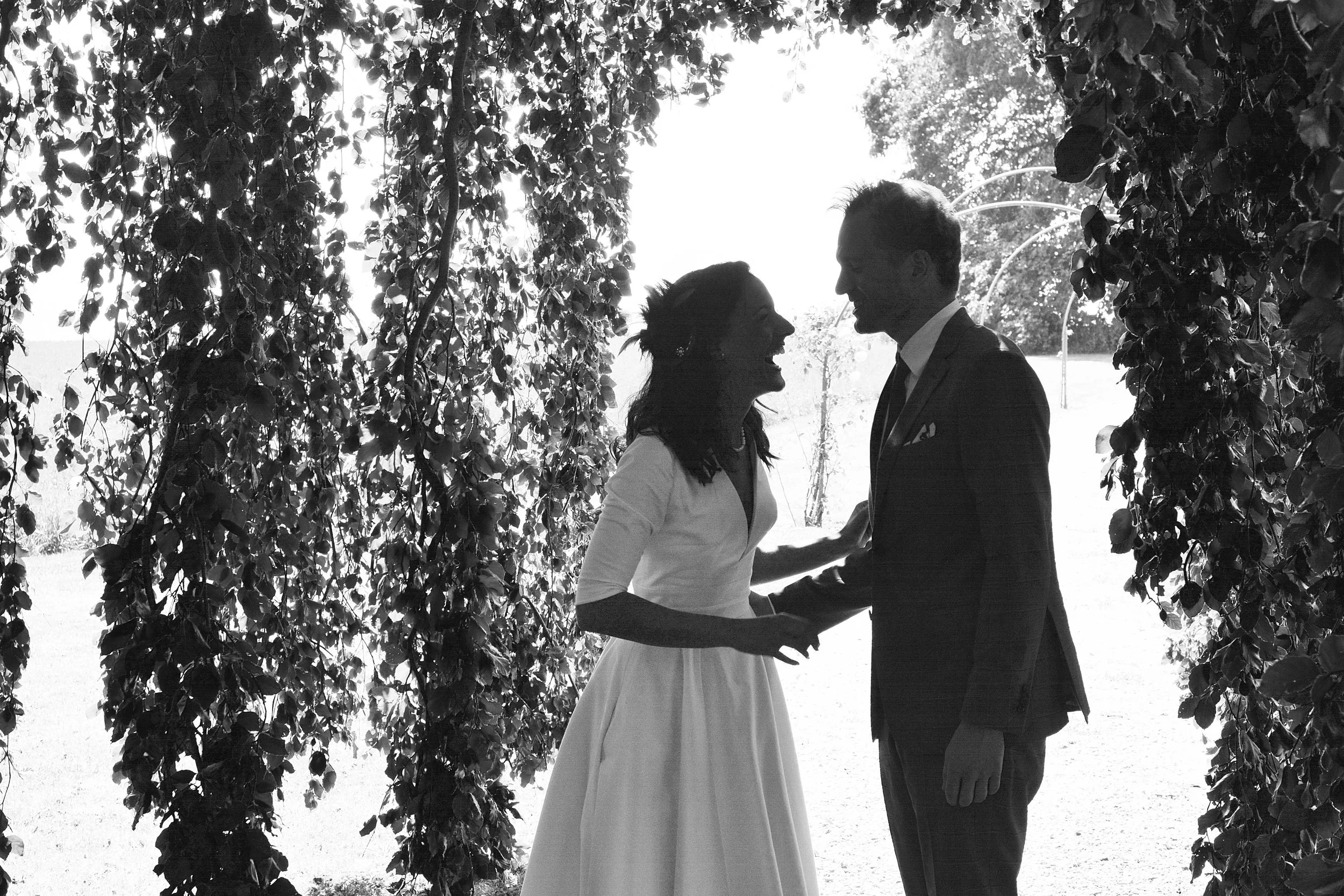 Un couple en vêtements de mariage se tenant par les mains sous une arche de branches et de feuilles, en plein air, en noir et blanc.
