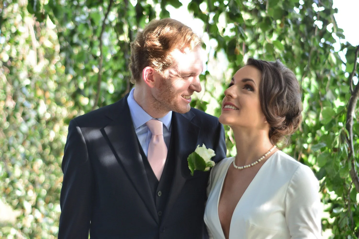 Un couple marié regarde l'un l'autre dans un environnement verdoyant, avec des feuilles en arrière-plan. La femme porte une robe blanche et un collier de perles, et l'homme porte un costume noir avec une cravate rose et une boutonnière blanche.