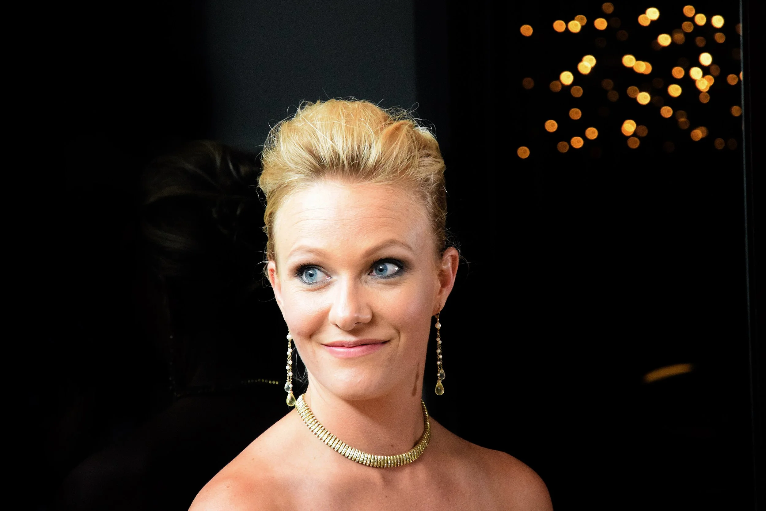 Portrait d'une femme aux cheveux blonds, portant des bijoux dorés, souriant légèrement.