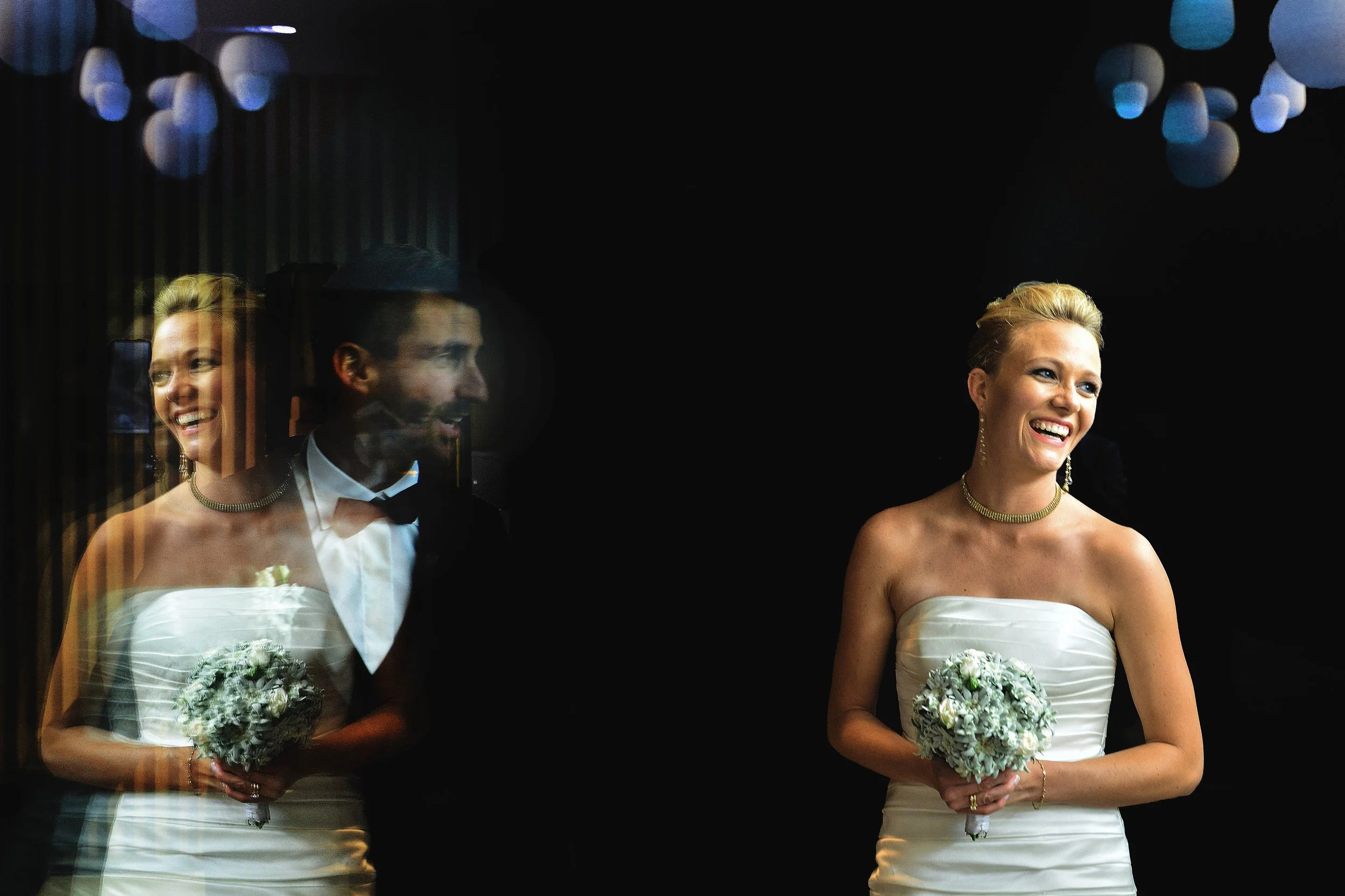Une mariée souriante portant une robe blanche, tenant un bouquet de fleurs, en compagnie d'un homme portant un costume avec nœud papillon, avec une moitié de son visage visible en apparaissant comme un reflet ou à travers une surface vitrée.