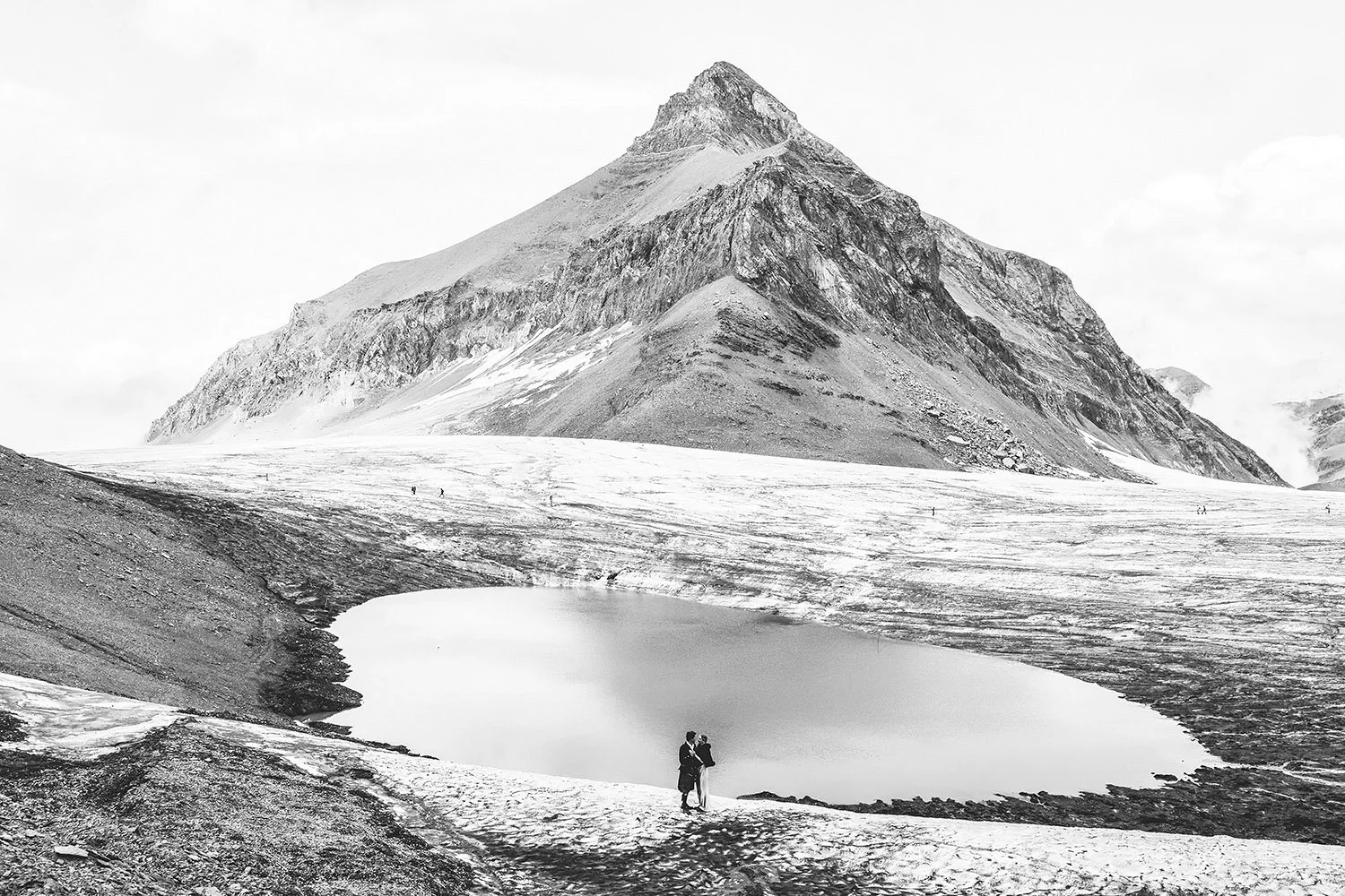 Deux personnes se tenant par la taille près d'un lac de montagne, avec un grand pic rocheux en arrière-plan, en noir et blanc.