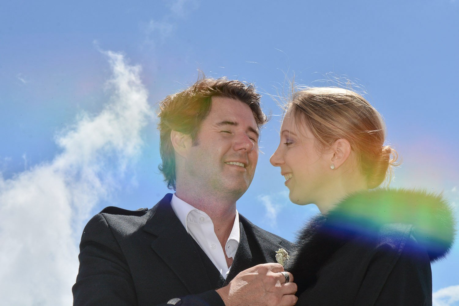 Un couple souriant lors de leur mariage, échangeant un geste d'affection sous un ciel bleu avec quelques nuages.
