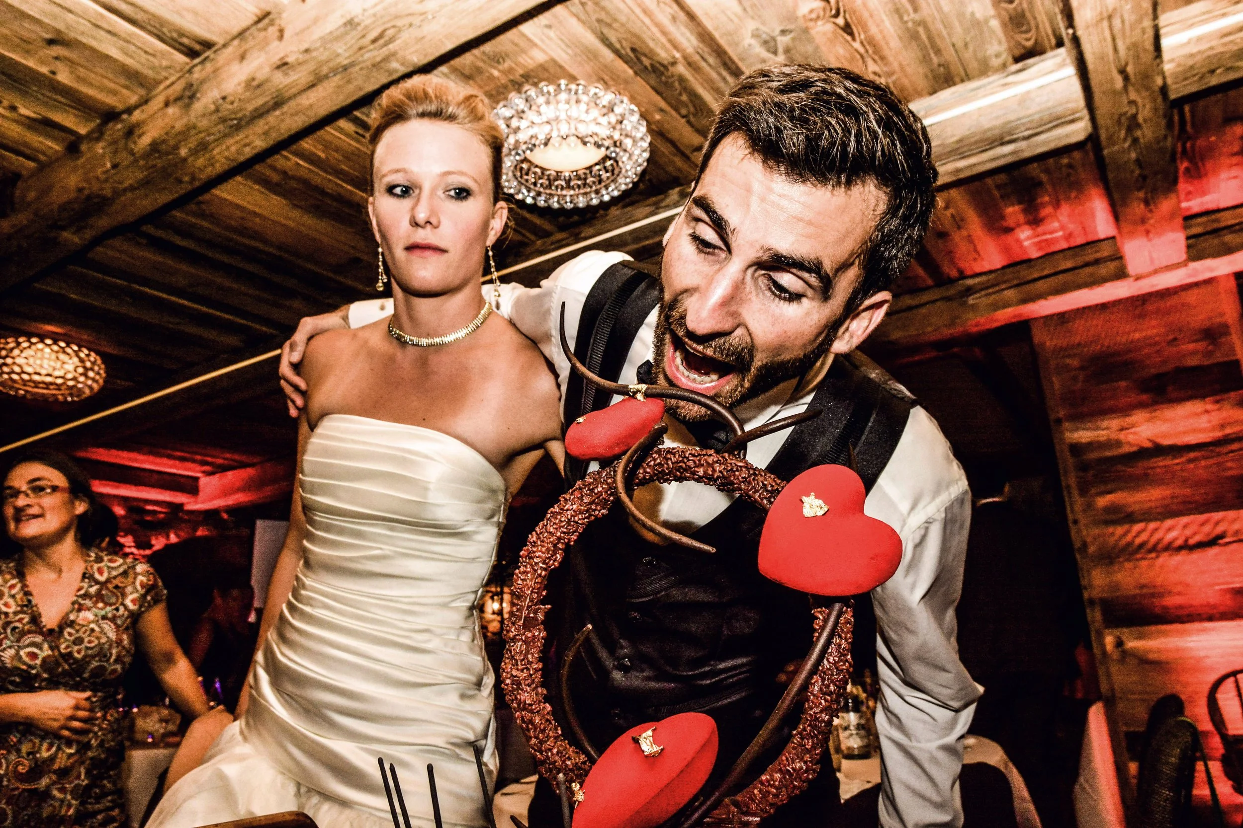 Une homme et une femme dans une pièce en bois, célébrant avec des décorations de fête, l'homme porte des cœurs décoratifs et semble jouer ou manger un objet.