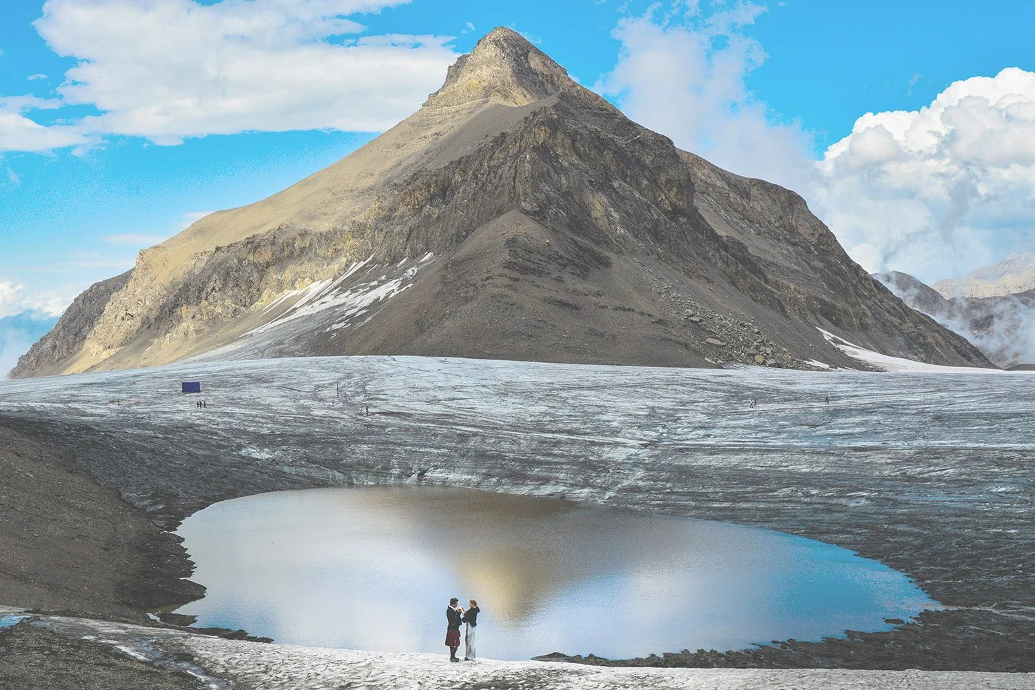 Deux personnes se tiennent près d’un lac de glacier au pied d’une montagne rocheuse, avec un ciel bleu et des nuages en arrière-plan.