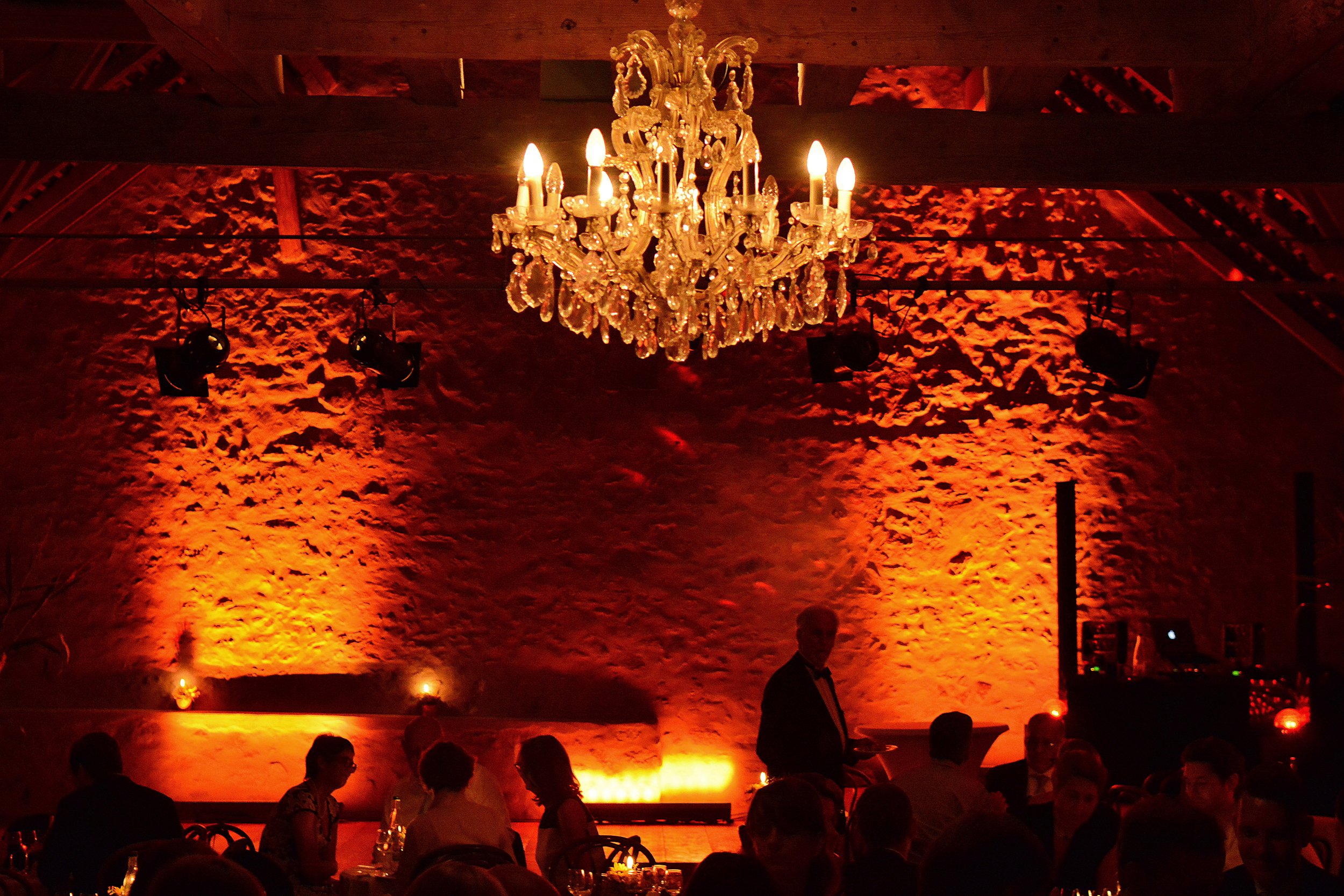 Une salle de banquet élégante avec un chandelier brillant suspendu au plafond, une ambiance chaleureuse avec des lumières oranges sur un mur en pierre, des personnes assises autour de tables, un homme en smoking tenant un plateau.