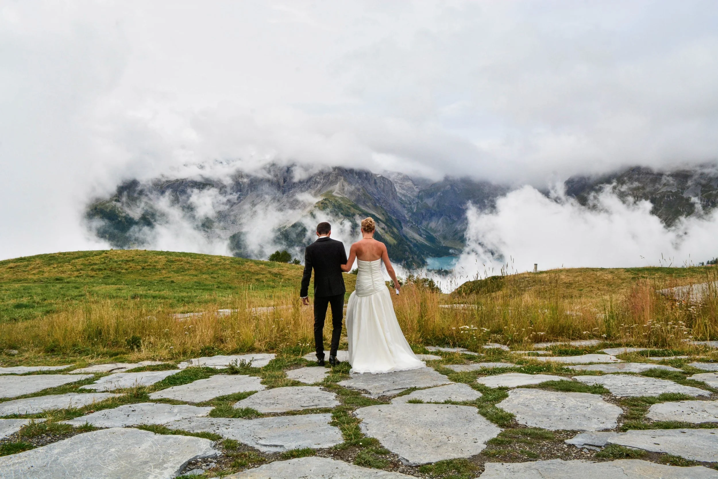 Un couple en tenue de mariage marche sur un chemin pavé vers un paysage de montagnes brumeuses.