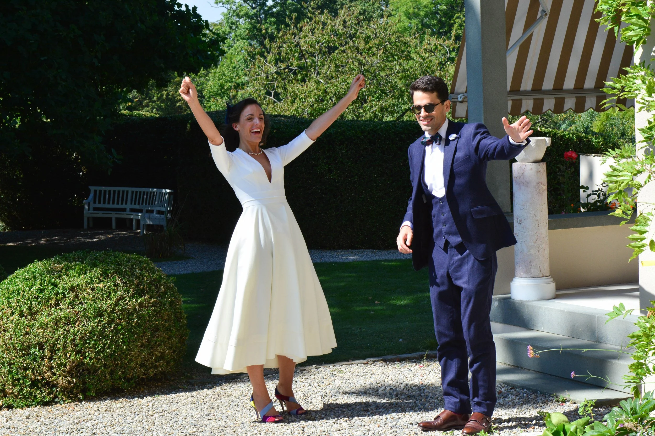Un couple habillé dans des vêtements de mariage, la femme en robe blanche et le homme en costume bleu marine, célébrant une occasion heureuse dans un jardin ensoleillé.