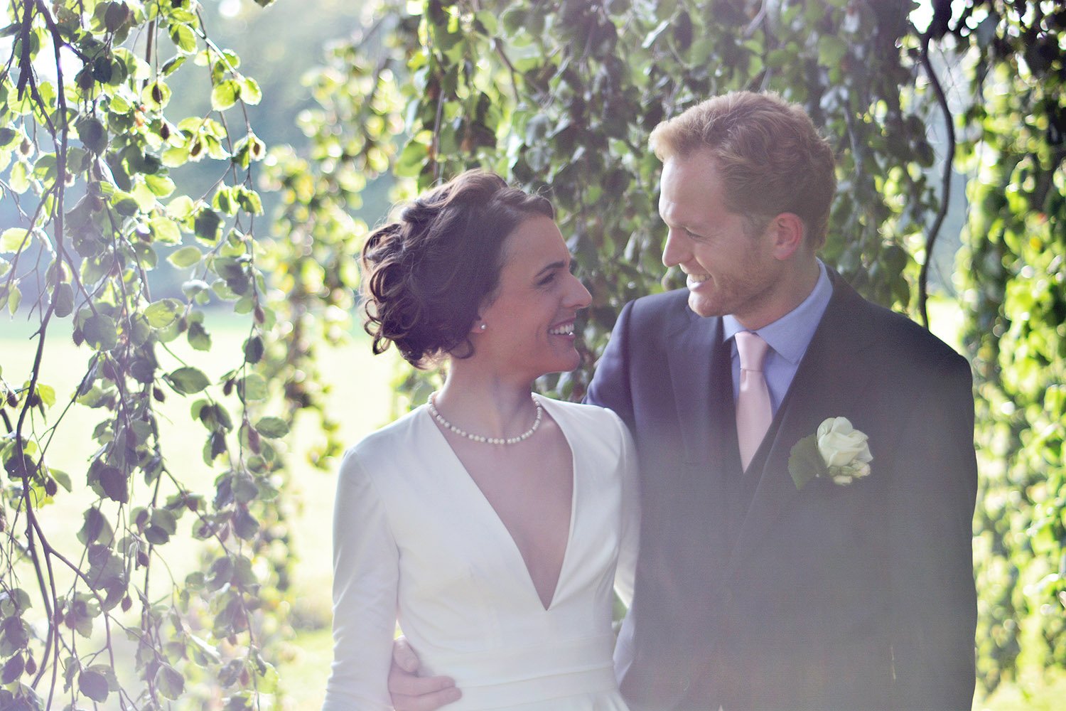 Un couple en costume de mariage, la femme en robe blanche et le homme en costume sombre avec une boutonnière, partage un moment joyeux dans un jardin entouré de feuillage vert.