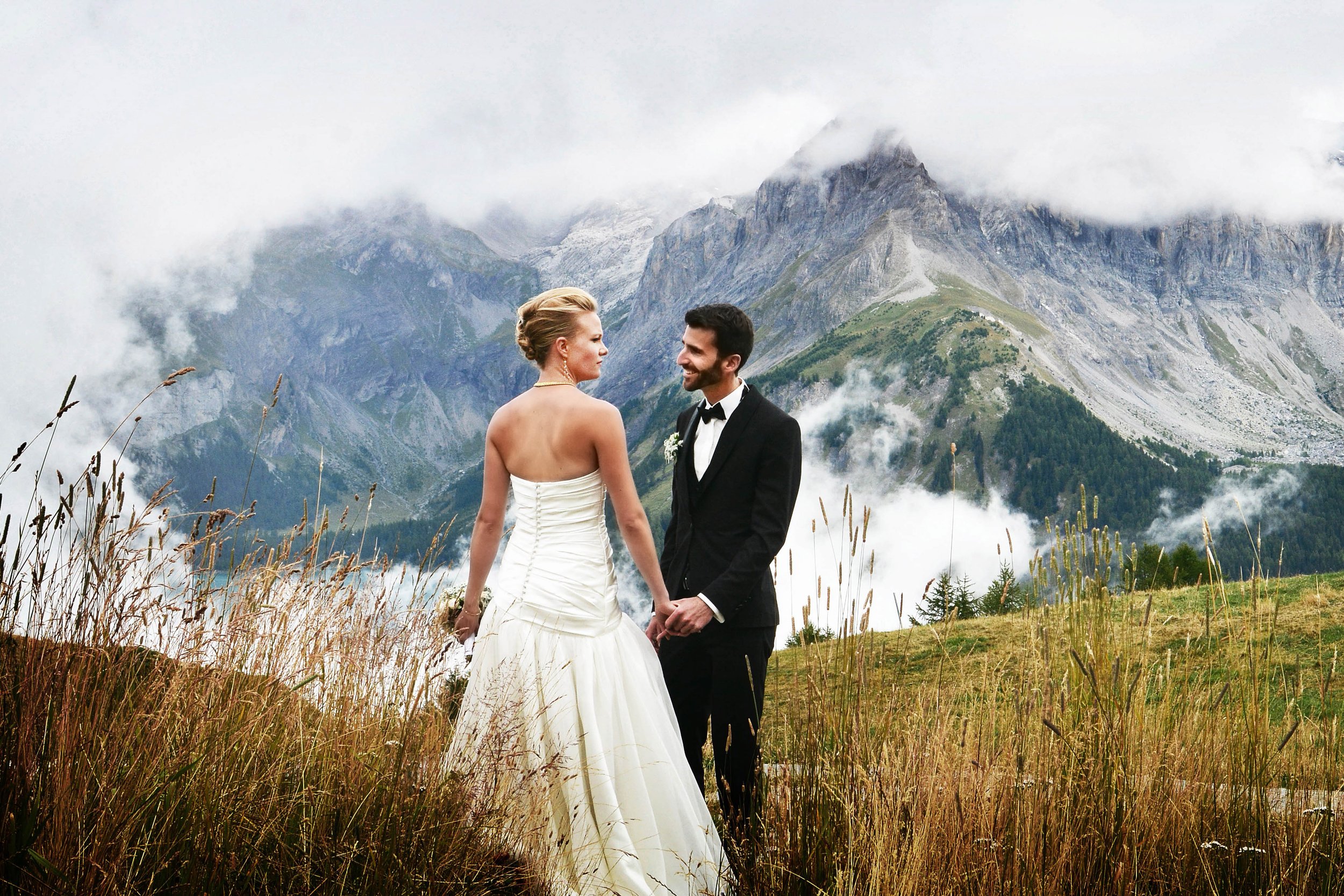 Un couple de mariés dans un paysage montagneux, la femme en robe blanche et l'homme en costume noir, se tenant par la main.