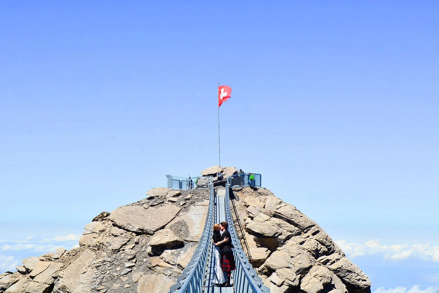 Deux personnes embrassent sur un pont suspendu au sommet d'une montagne rocheuse avec un drapeau suisse en haut.