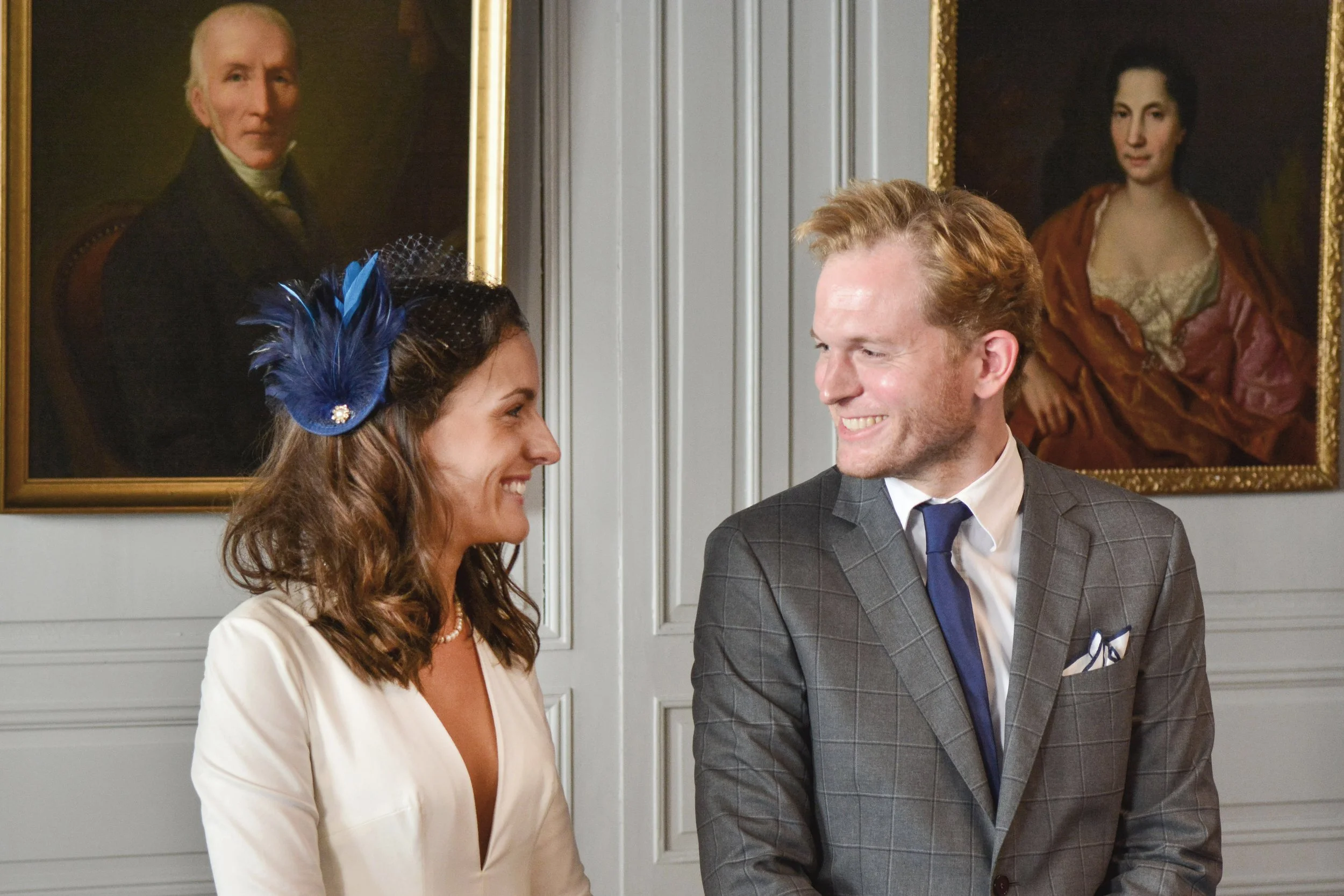 Un homme et une femme souriants regardant chacun l'autre, dans une pièce avec deux portraits en arrière-plan.