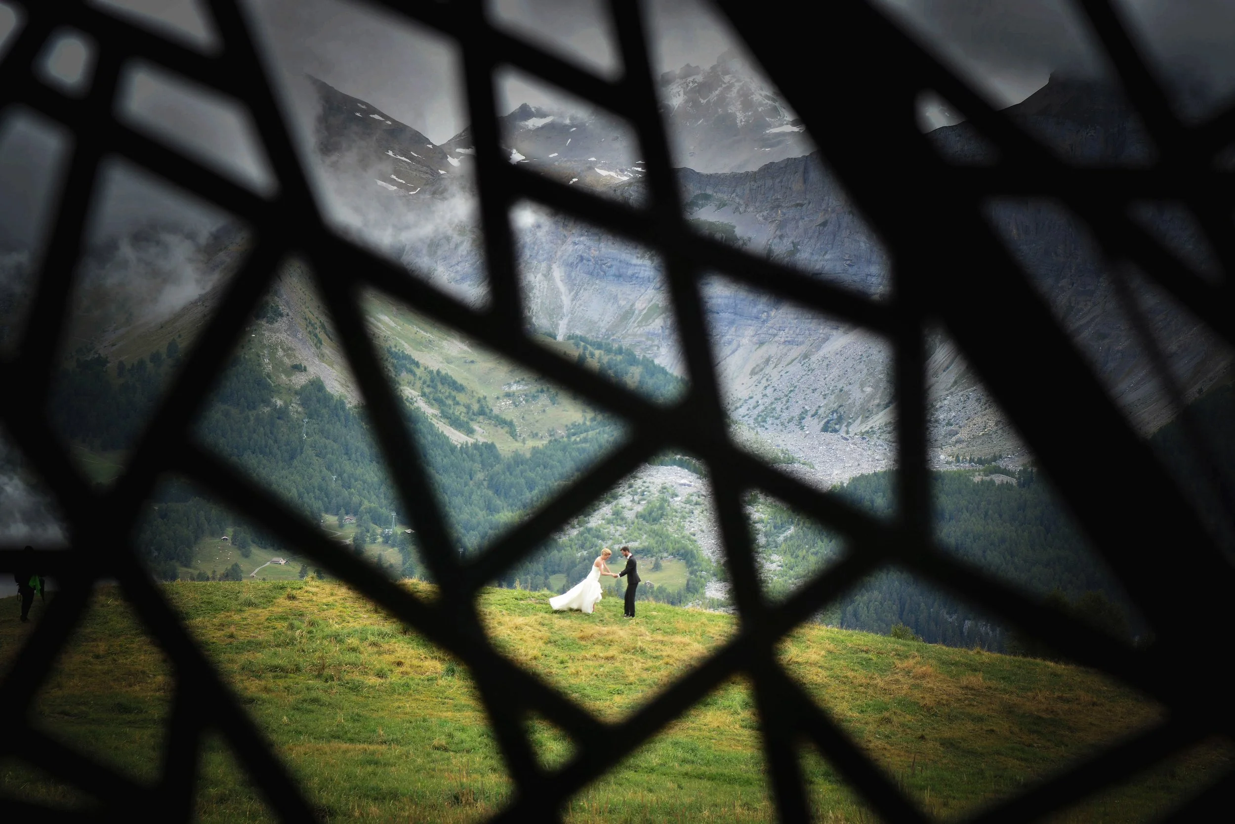 Un couple en vêtements de mariage, se tenant la main dans un paysage de montagnes verdoyantes, vue partielle à travers une structure en bois ou métal.