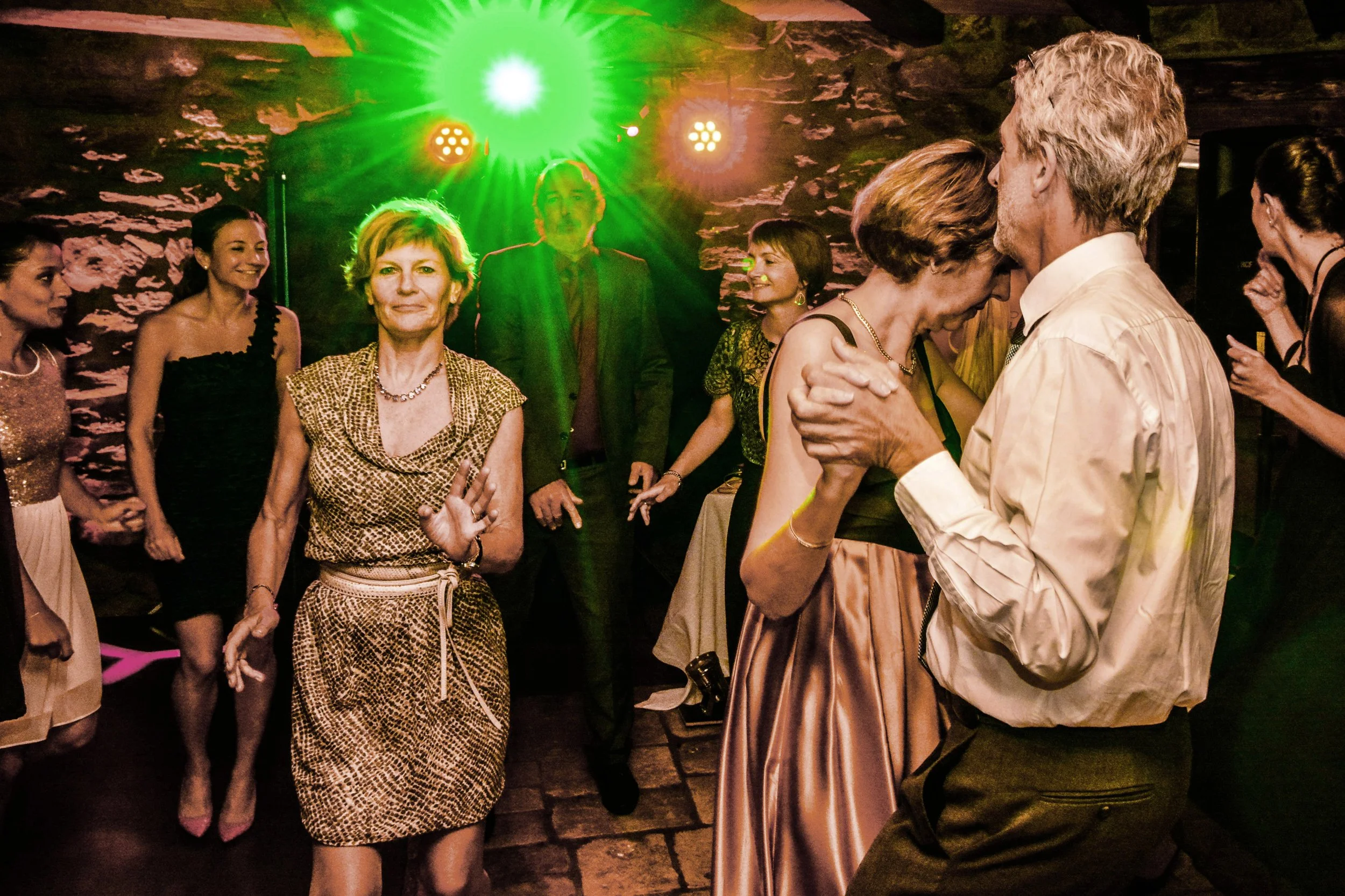 Des personnes dansent et s'amusent lors d'une fête ou d'un mariage dans une pièce avec une ambiance lumineuse verte et rose.